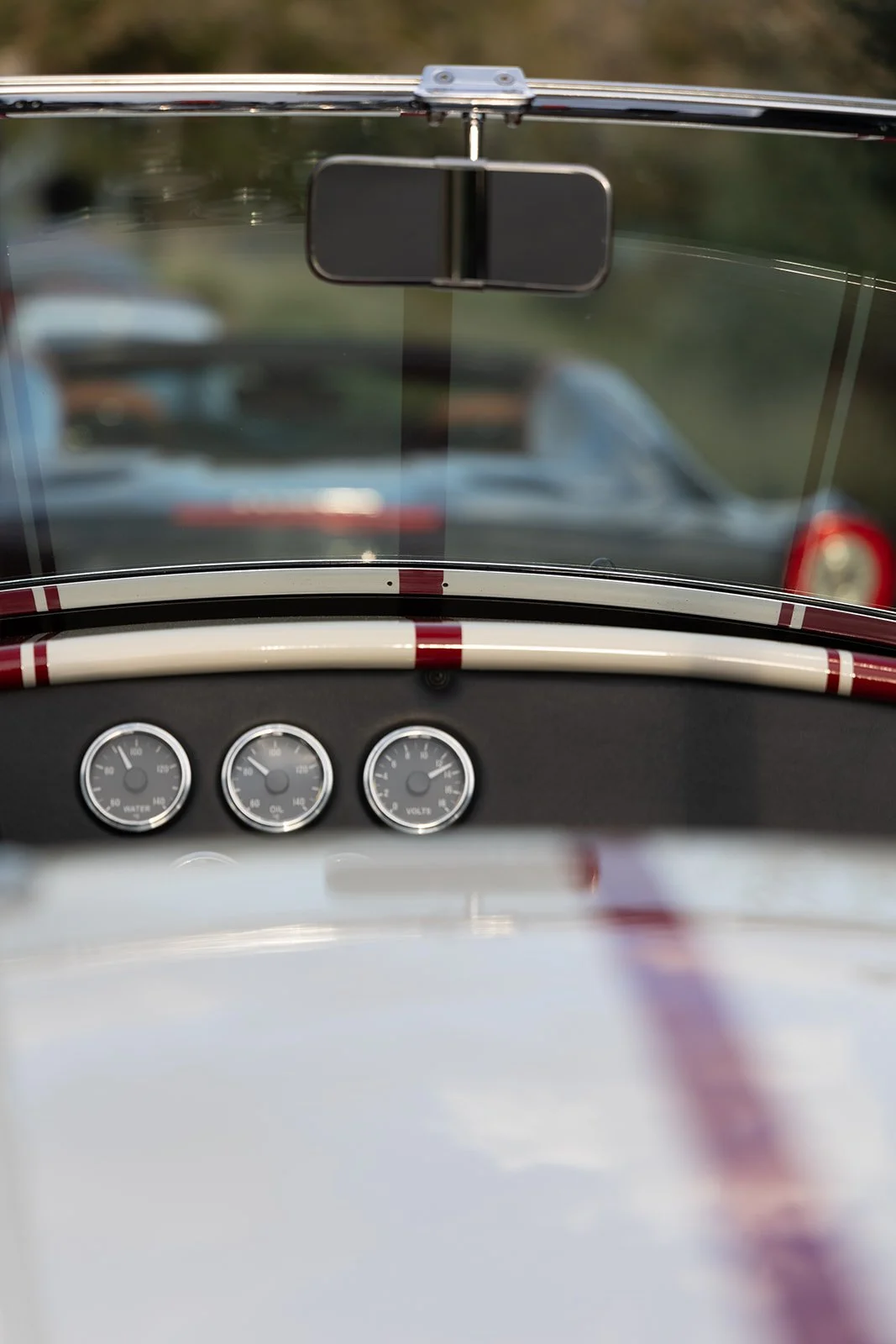 Dashboard of a vintage car with three gauges for water, oil, and voltage, and rearview mirror, with a blurred background of parked cars.