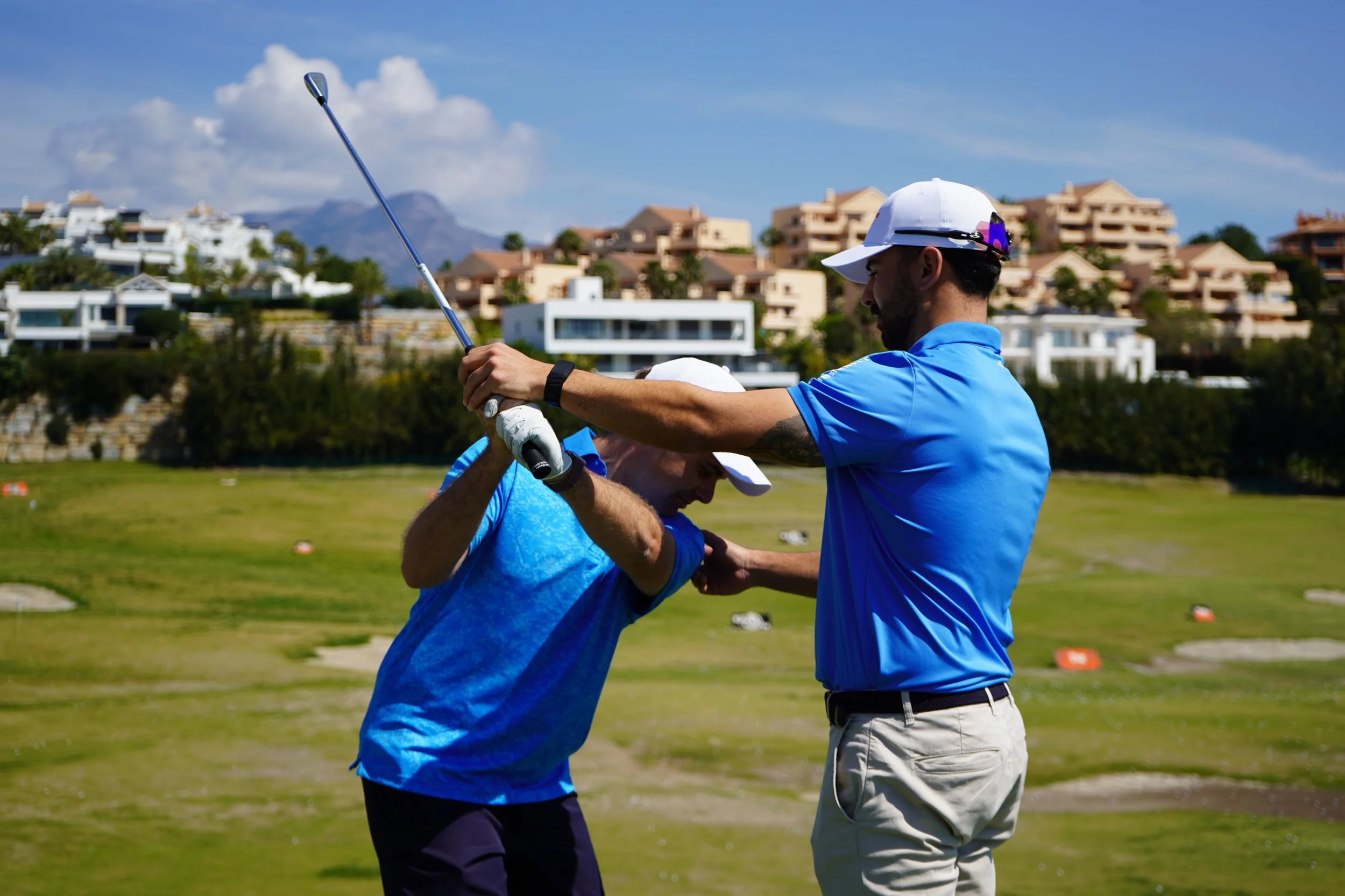 A man teaching another man how to golf swing on a golf course with houses in the background.