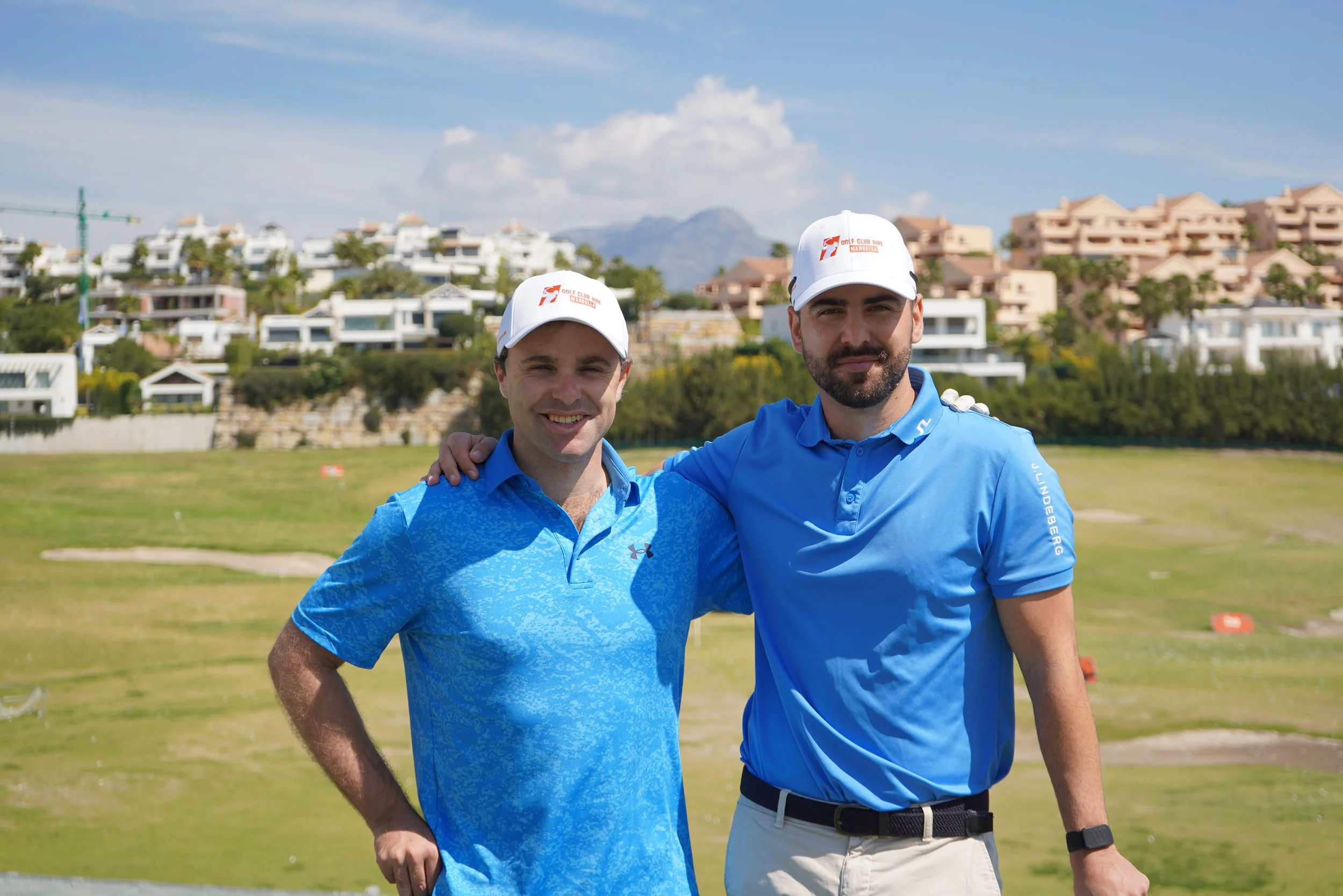 Two men in blue shirts and white caps standing together on a golf course with residential buildings and mountains in the background.