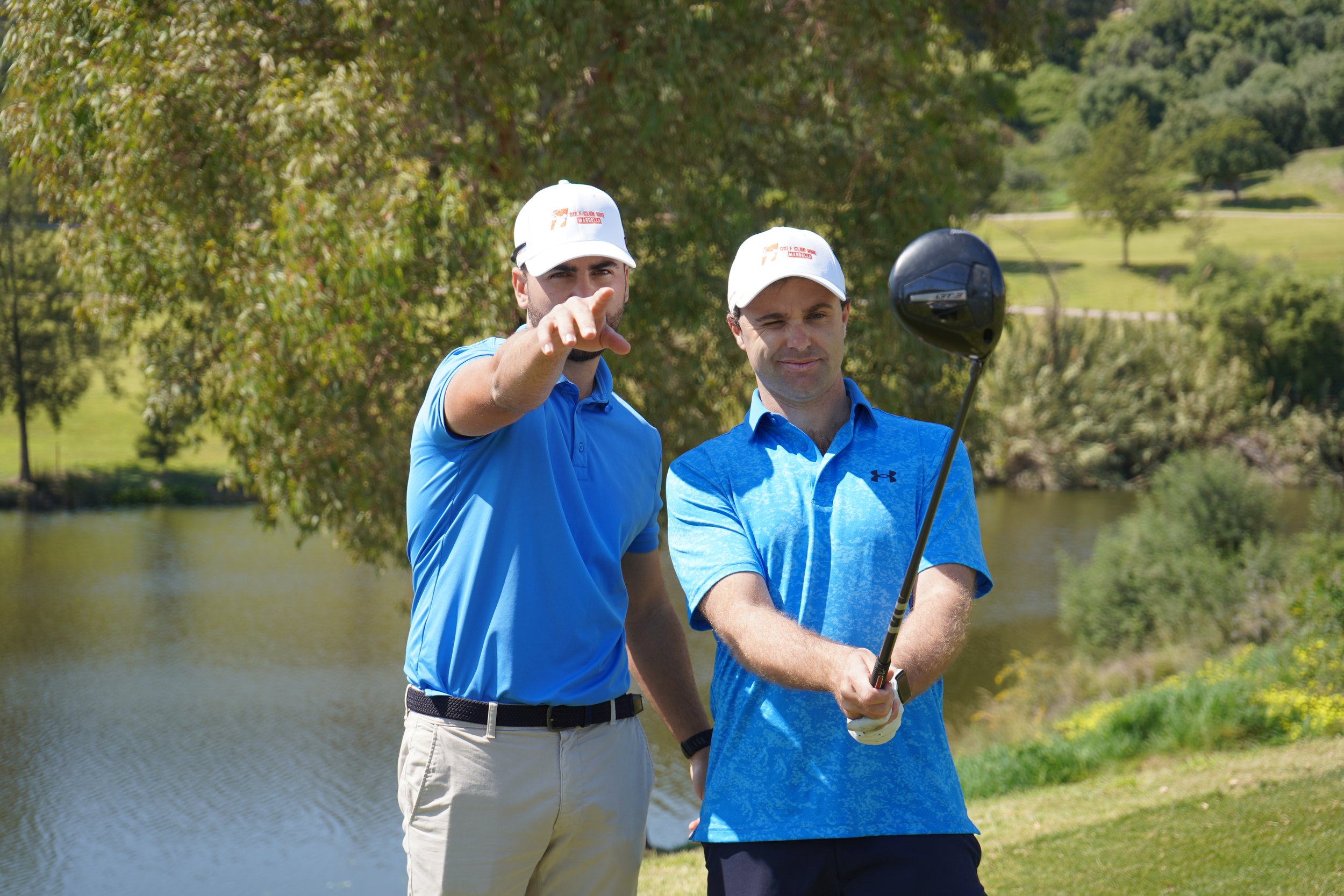Two men in golf attire standing on a golf course near a body of water, with trees and golf course landscape in the background. One man is pointing while the other is holding a golf club.