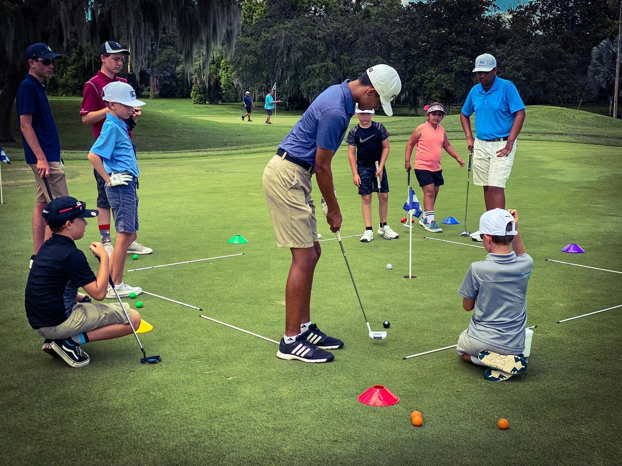 A group of children and a coach practicing golf on a putting green surrounded by trees.