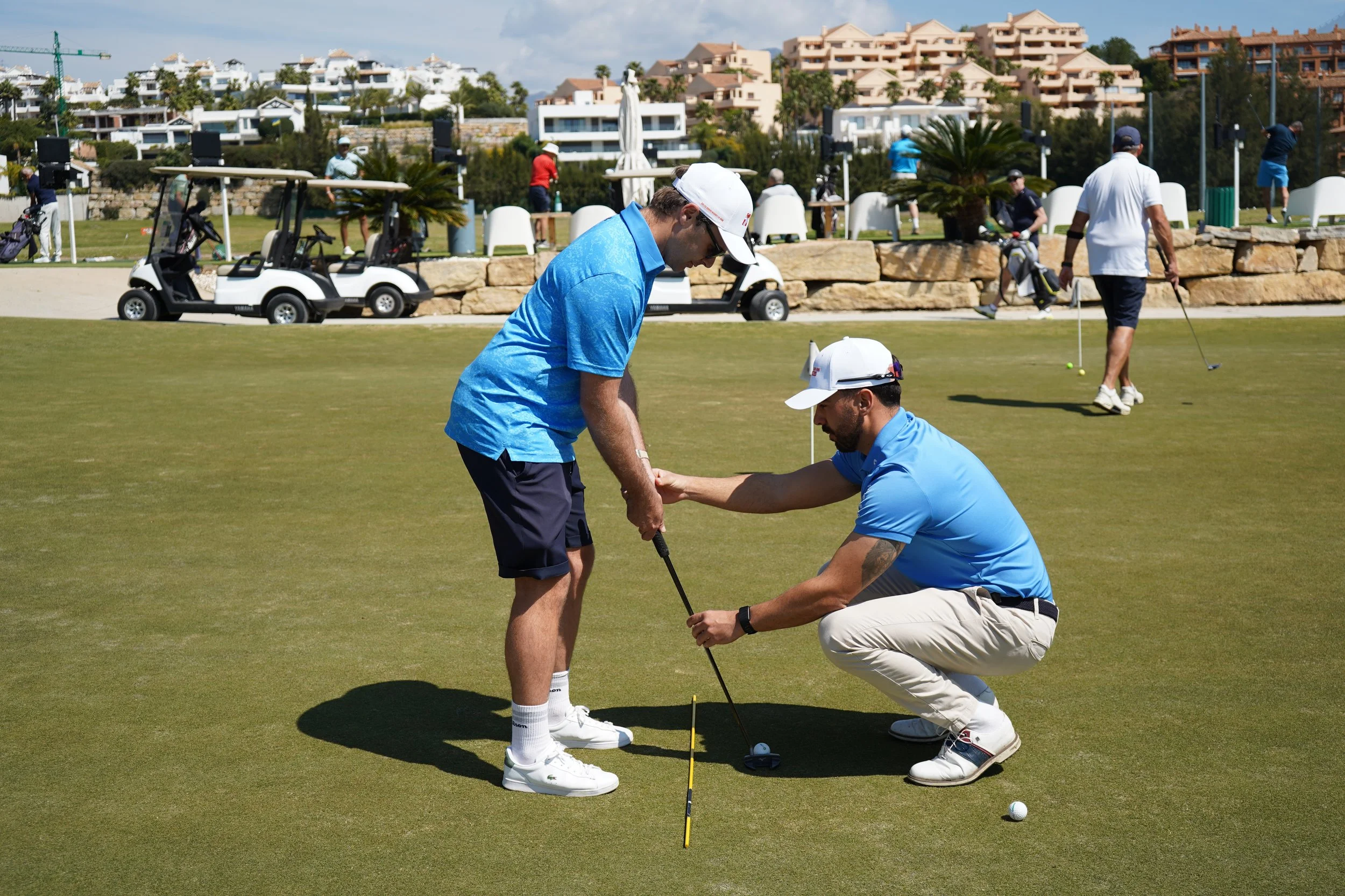 A golf instructor helping a young man with his golf stance and grip on the putting green at a golf course, with other players and golf carts in the background.