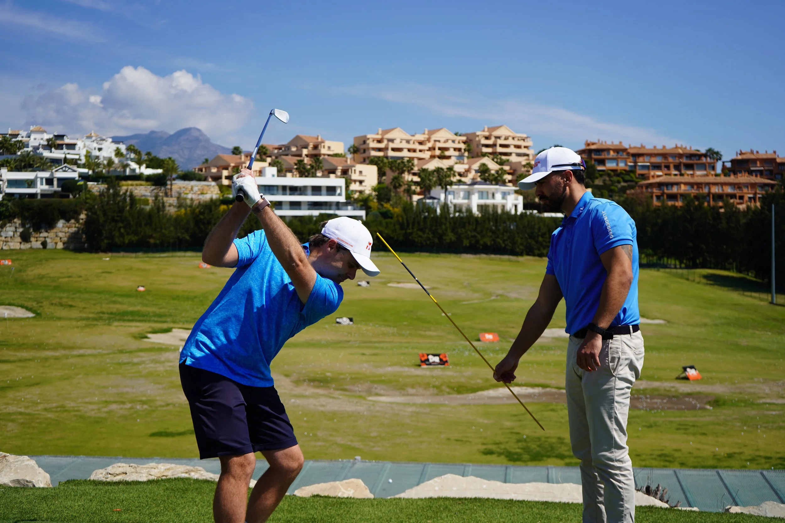 A man taking a golf swing with a golf club while another man, possibly a coach, stands beside him holding a golf tee on a golf course under a blue sky.