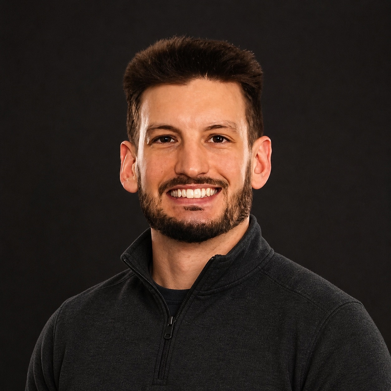 Headshot of a smiling man with a beard and short brown hair, wearing a dark zip-up sweater, against a black background.