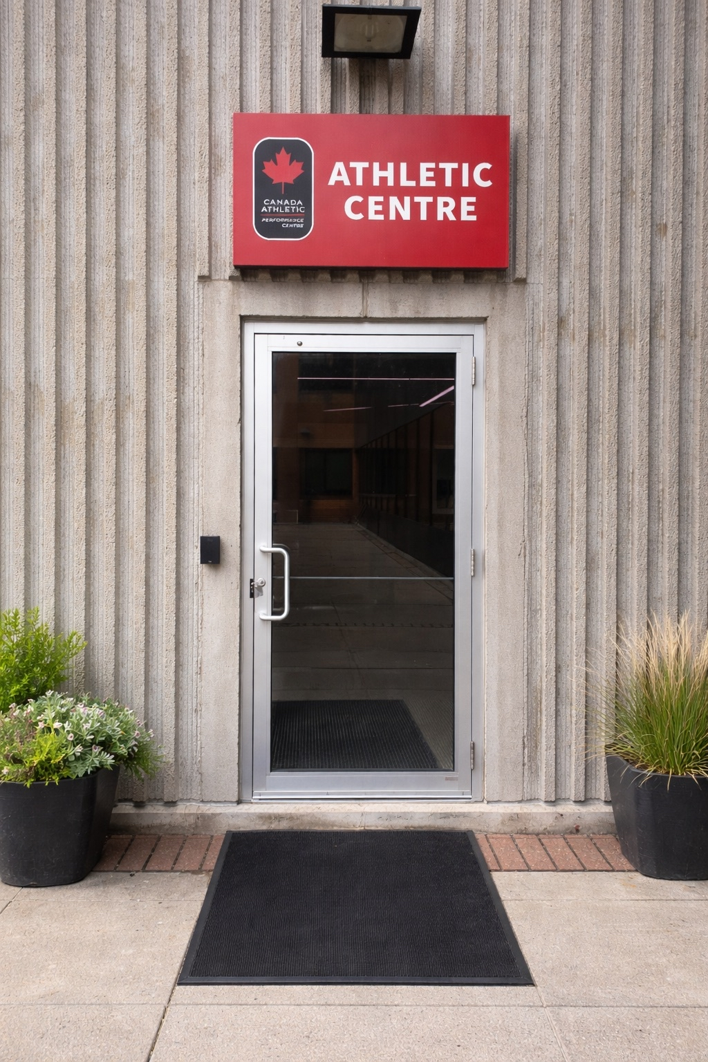 Exterior view of the entrance to a Canadian Athletic Centre with a glass door, potted plants on either side, and a red sign above that reads 'Athletic Centre' with a maple leaf logo.