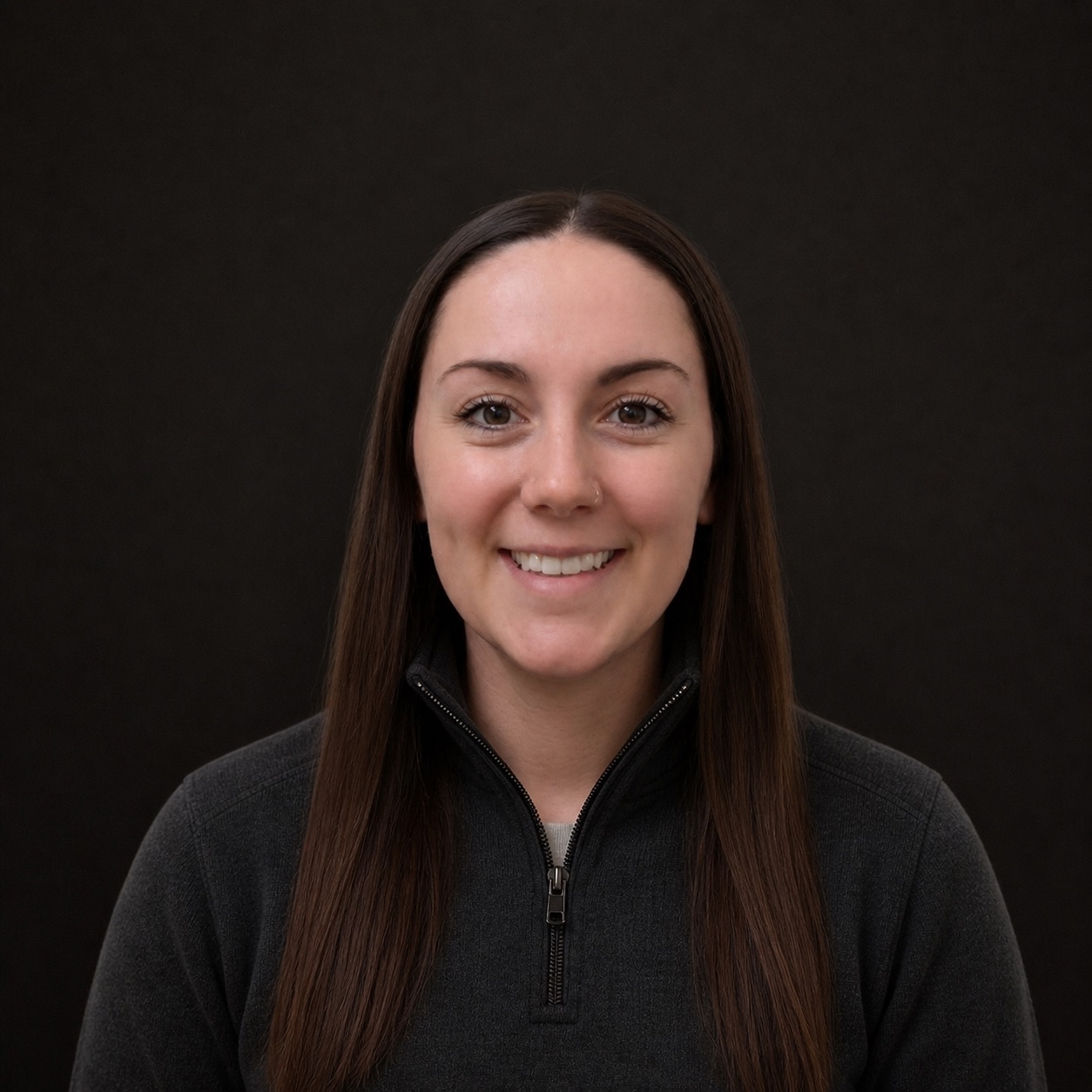 Portrait of a young woman with long brown hair, wearing a black zip-up jacket, smiling, against a dark background.