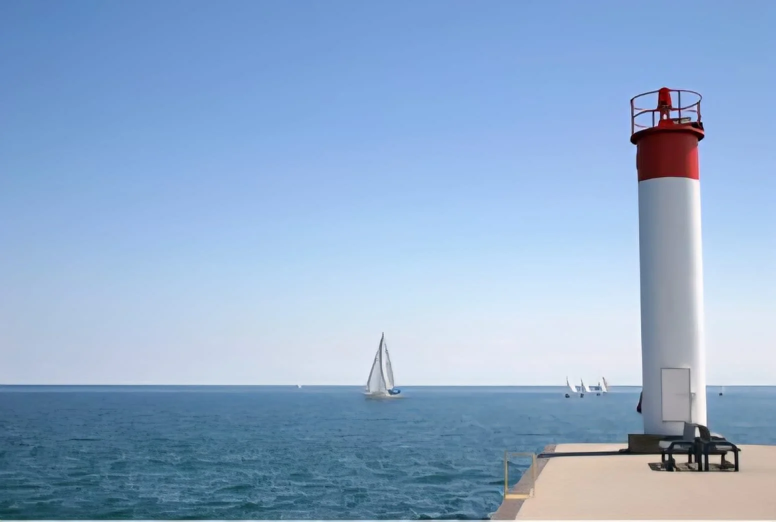 A lighthouse on a dock near the ocean with sailboats in the background and a clear blue sky.