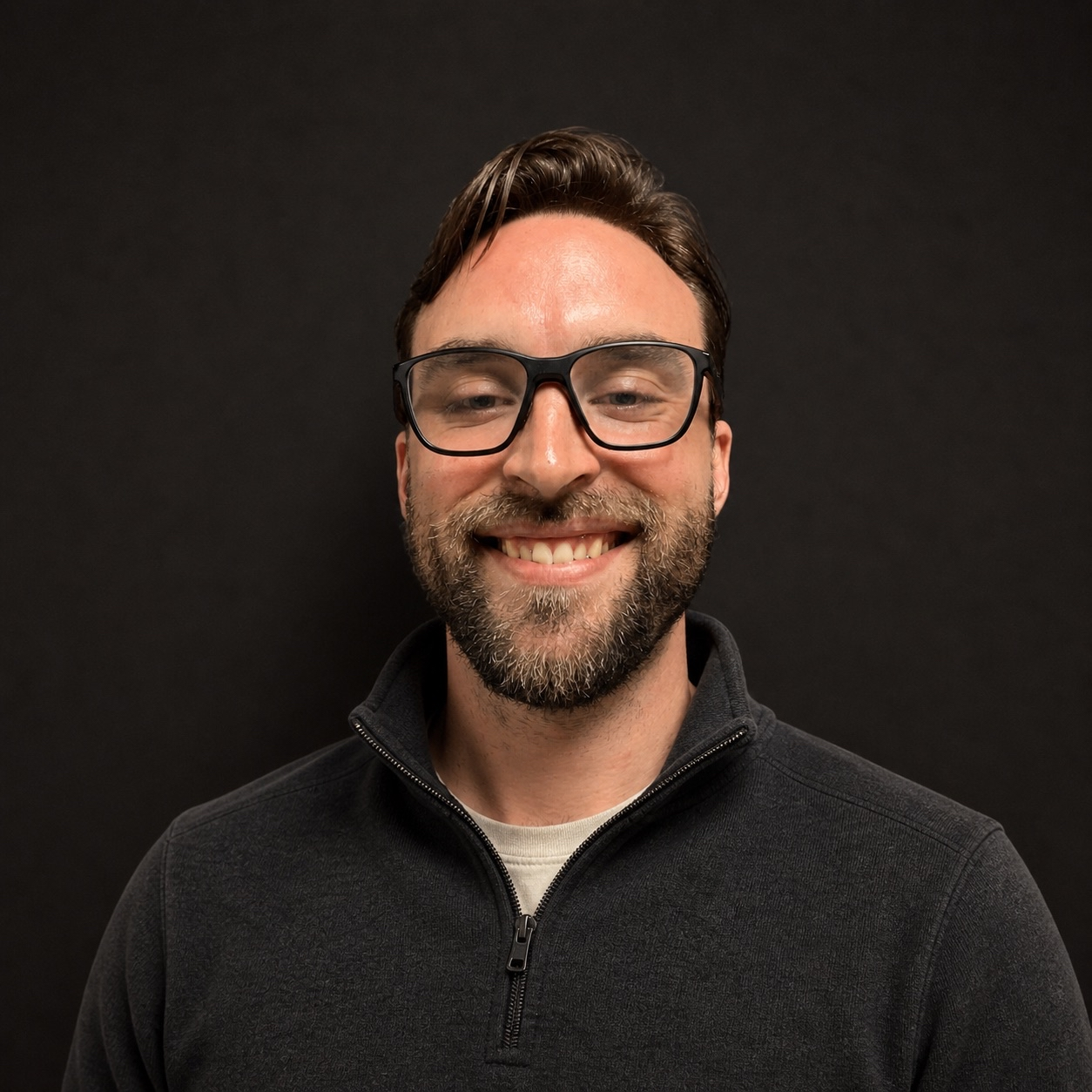 A man with glasses, beard, and mustache smiling against a plain black background.