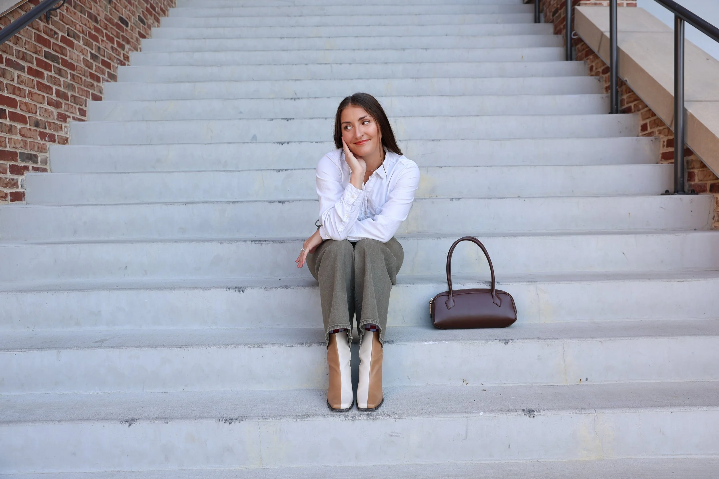 Jessica Clark sitting on white concrete steps with a brick wall behind her, wearing a white shirt, green pants, and white & brown boots, with a brown handbag placed next to her.