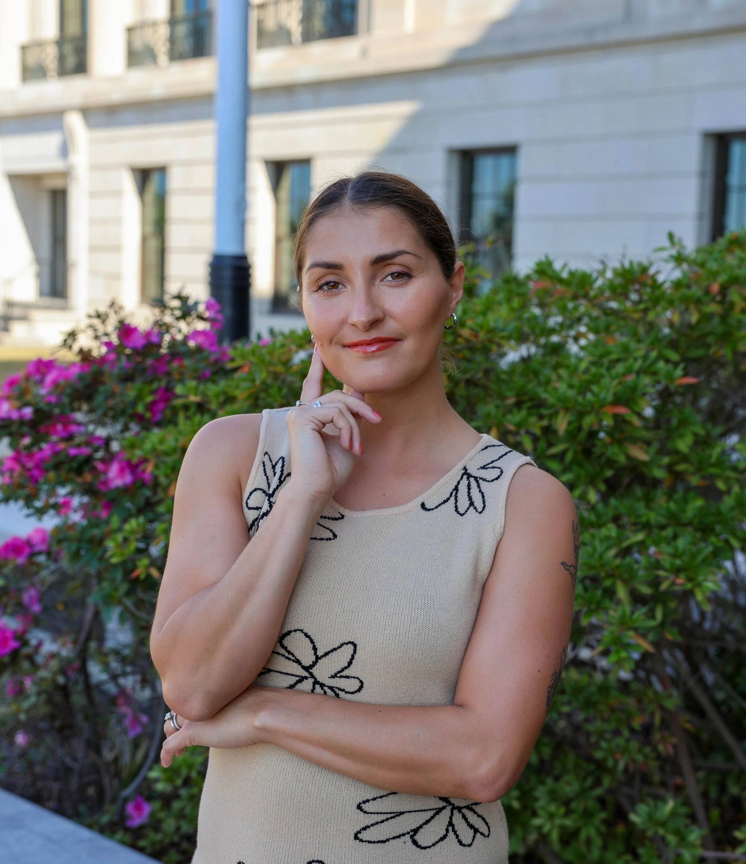 Jessica Clark standing outdoors in front of green bushes with pink flowers, wearing a beige sleeveless dress with black floral patterns, looking at the camera with a subtle smile.