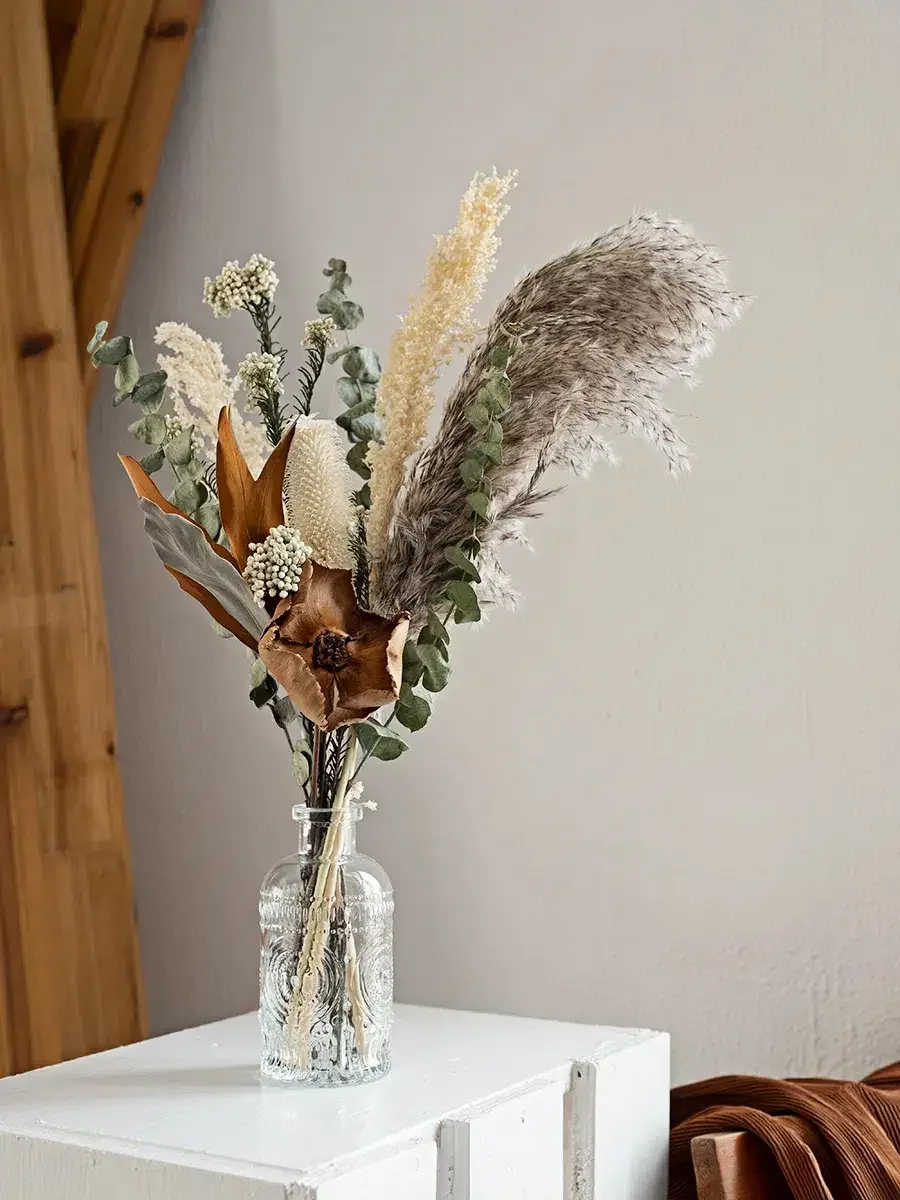 Dried floral arrangement in a textured glass vase on a white surface, with a wooden wall and a beige wall in the background.