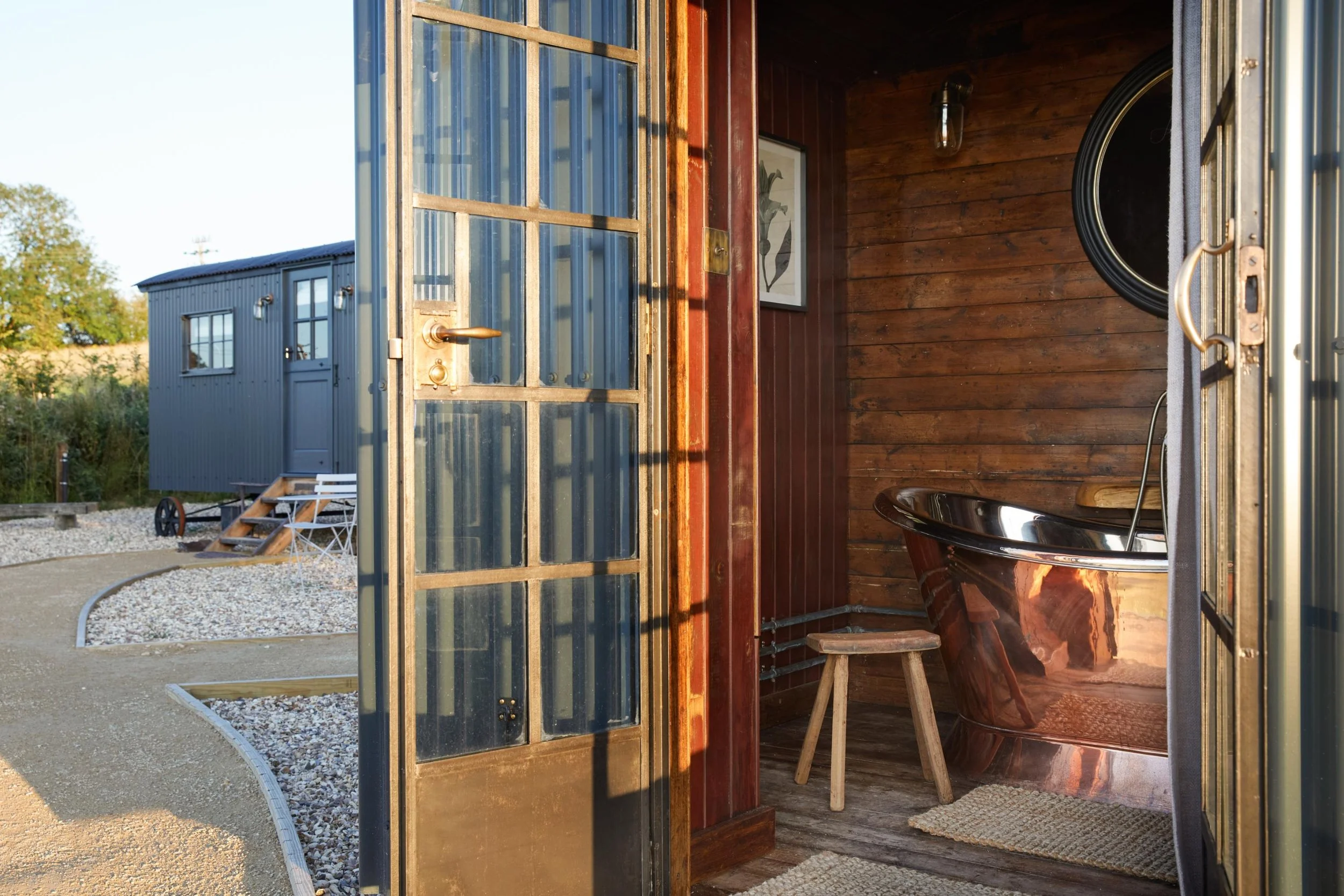 Interior view of a wooden sauna with a copper bathtub, a wooden stool, a framed botanical print, and an interior light fixture, with an open door leading outside.