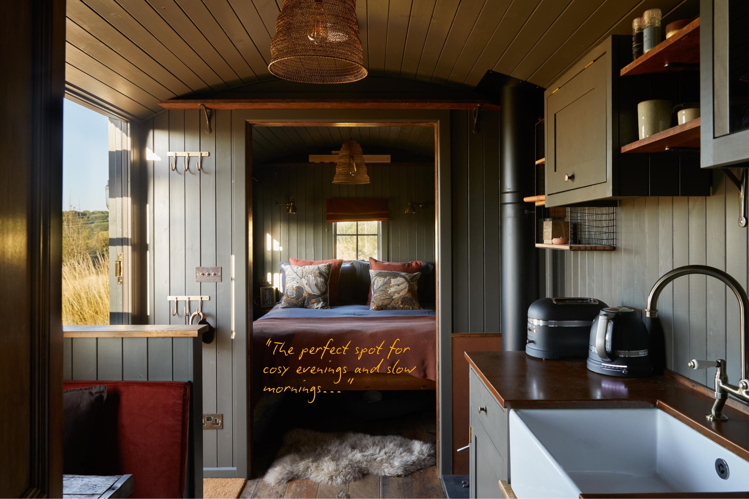 Interior of a cozy tiny home or cabin with a view into a bedroom. The bedroom has a bed with patterned pillows, small wooden shelves, and wall-mounted lamps. The room is lit by natural light coming through a window, and there are warm wooden ceiling 
