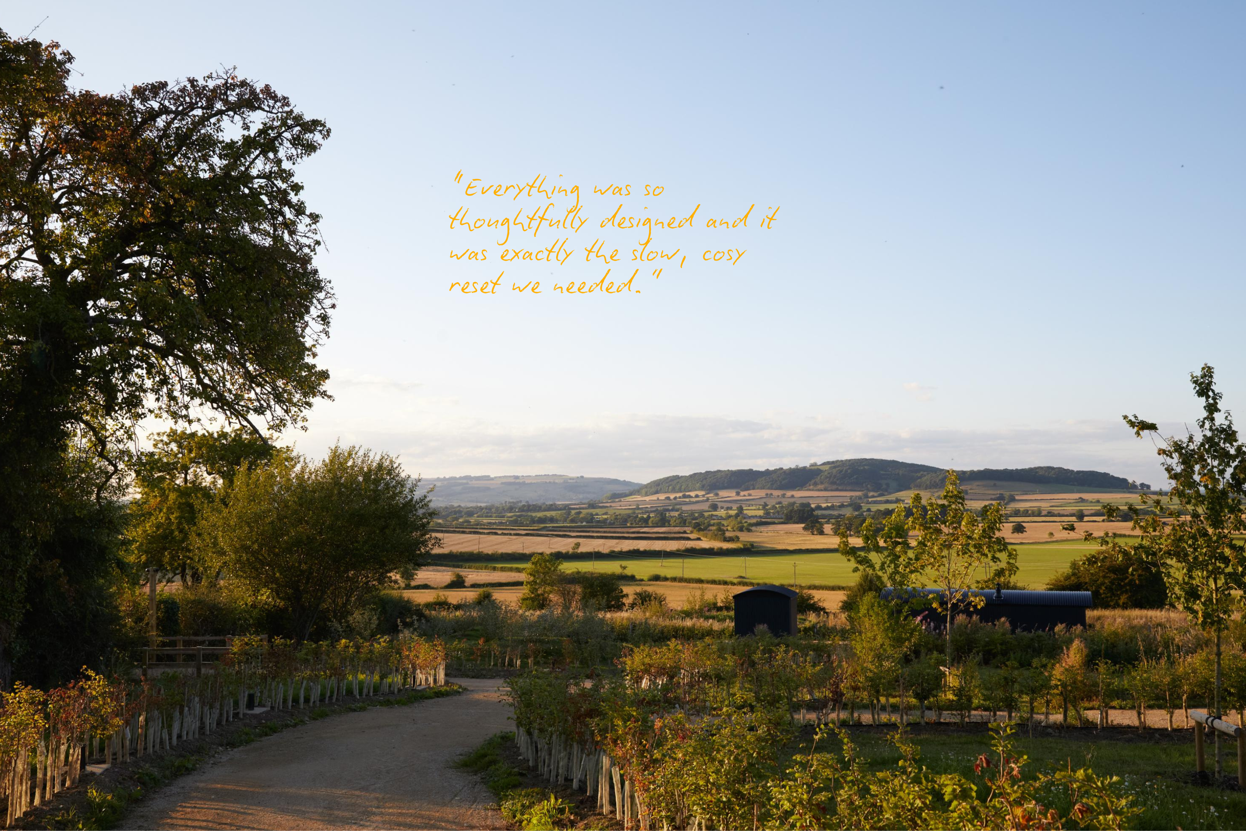 Scenic countryside landscape with a large tree on the left, a path in the foreground, fields and hills in the distance, and a quote in the sky reading 'Everything was so thoughtfully designed and it was exactly the slow, cosy reset we needed.'