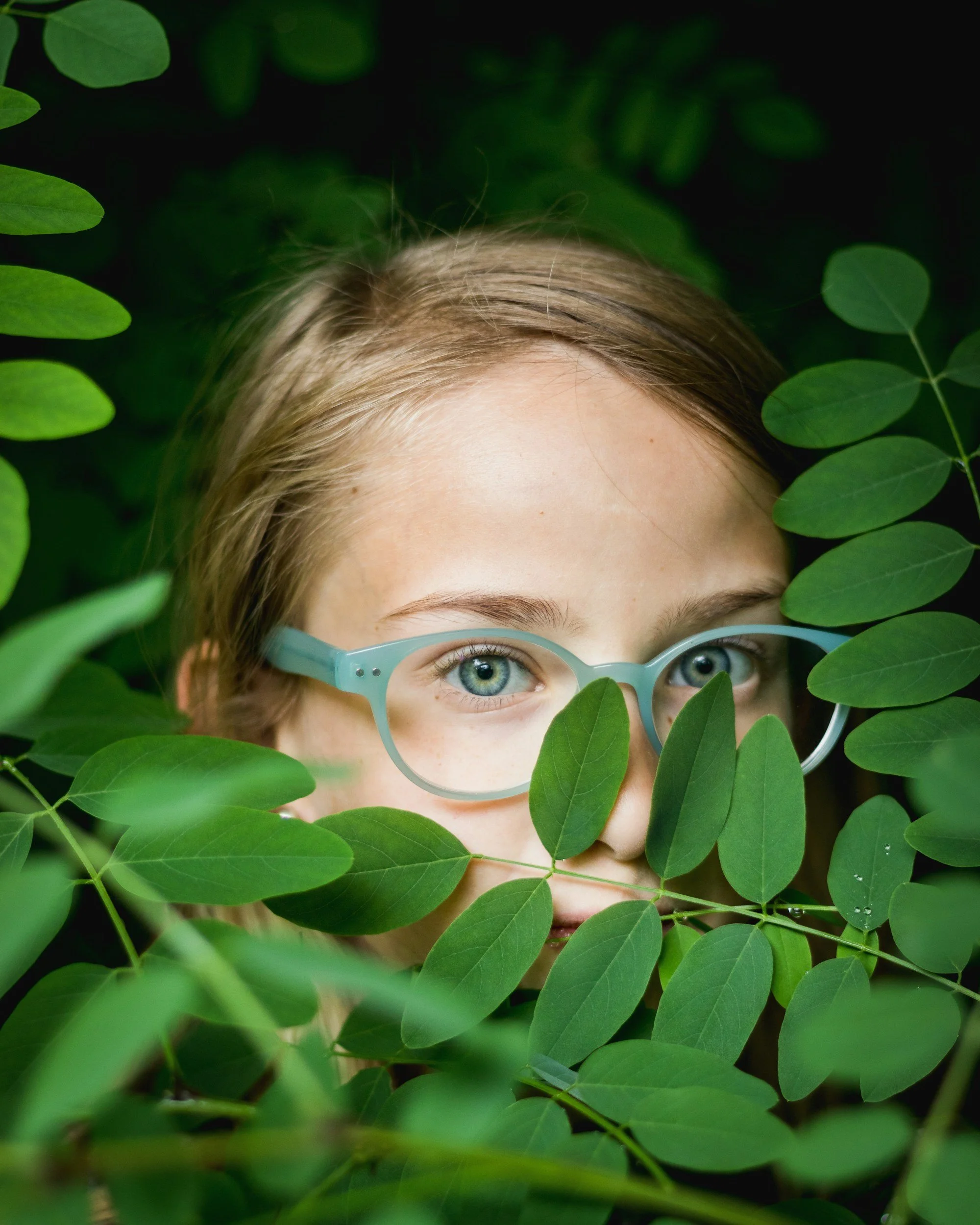 Close-up of a woman with blonde hair and blue eyes, wearing glasses, partially obscured by green leaves.