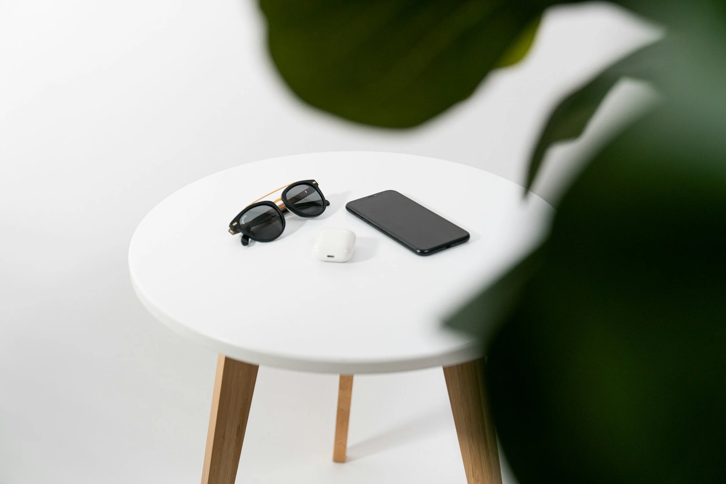 A white round table with wooden legs, on which are sunglasses, a smartphone, and wireless earbuds, partially obscured by green leaves.