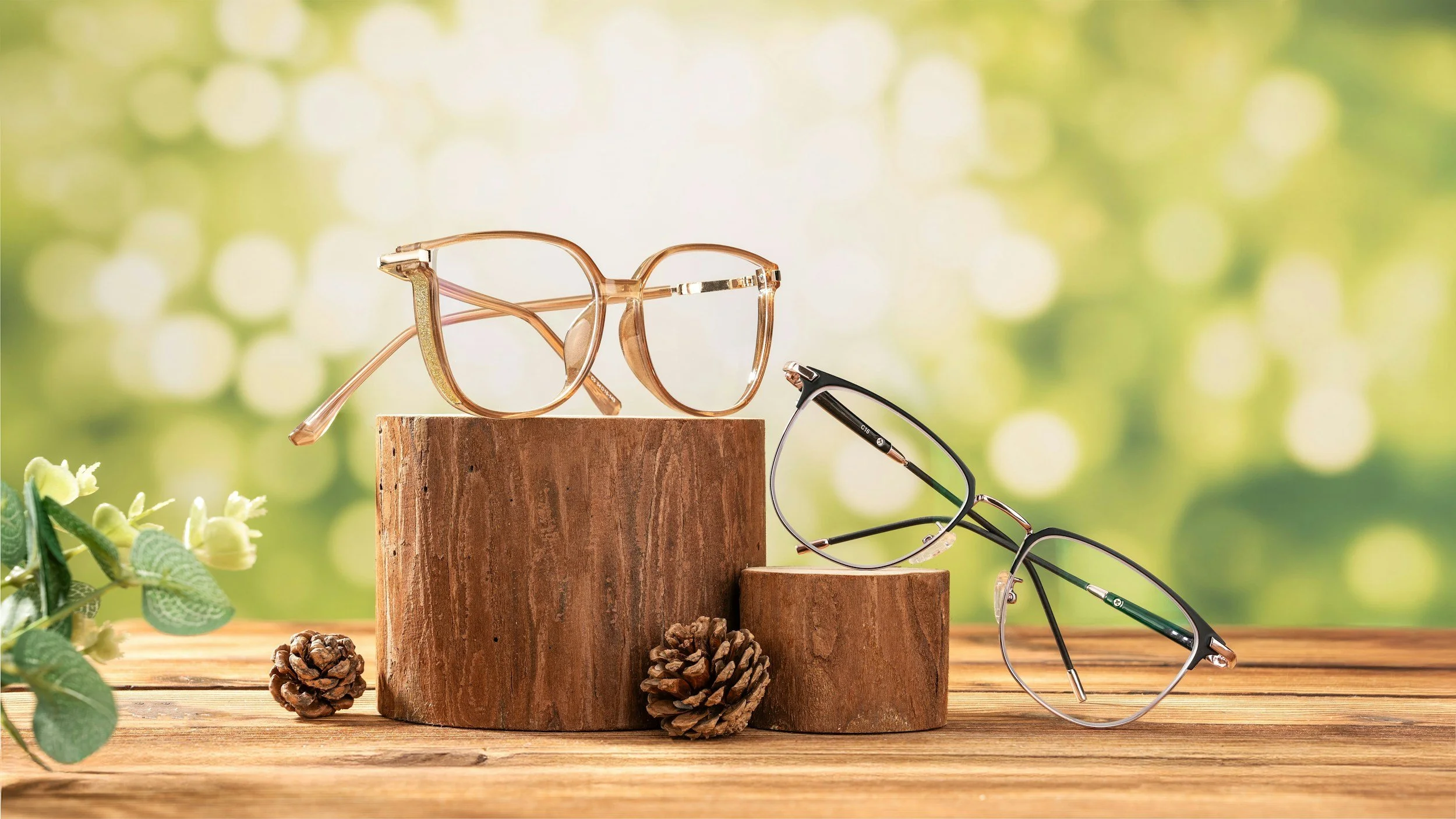 Three pairs of eyeglasses on wooden blocks with pinecones and greenery in the background.