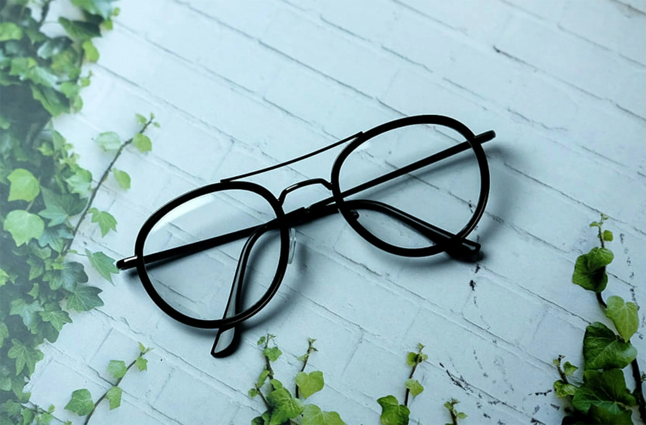 Black metal eyeglasses resting on a light gray wooden surface surrounded by green leaves.