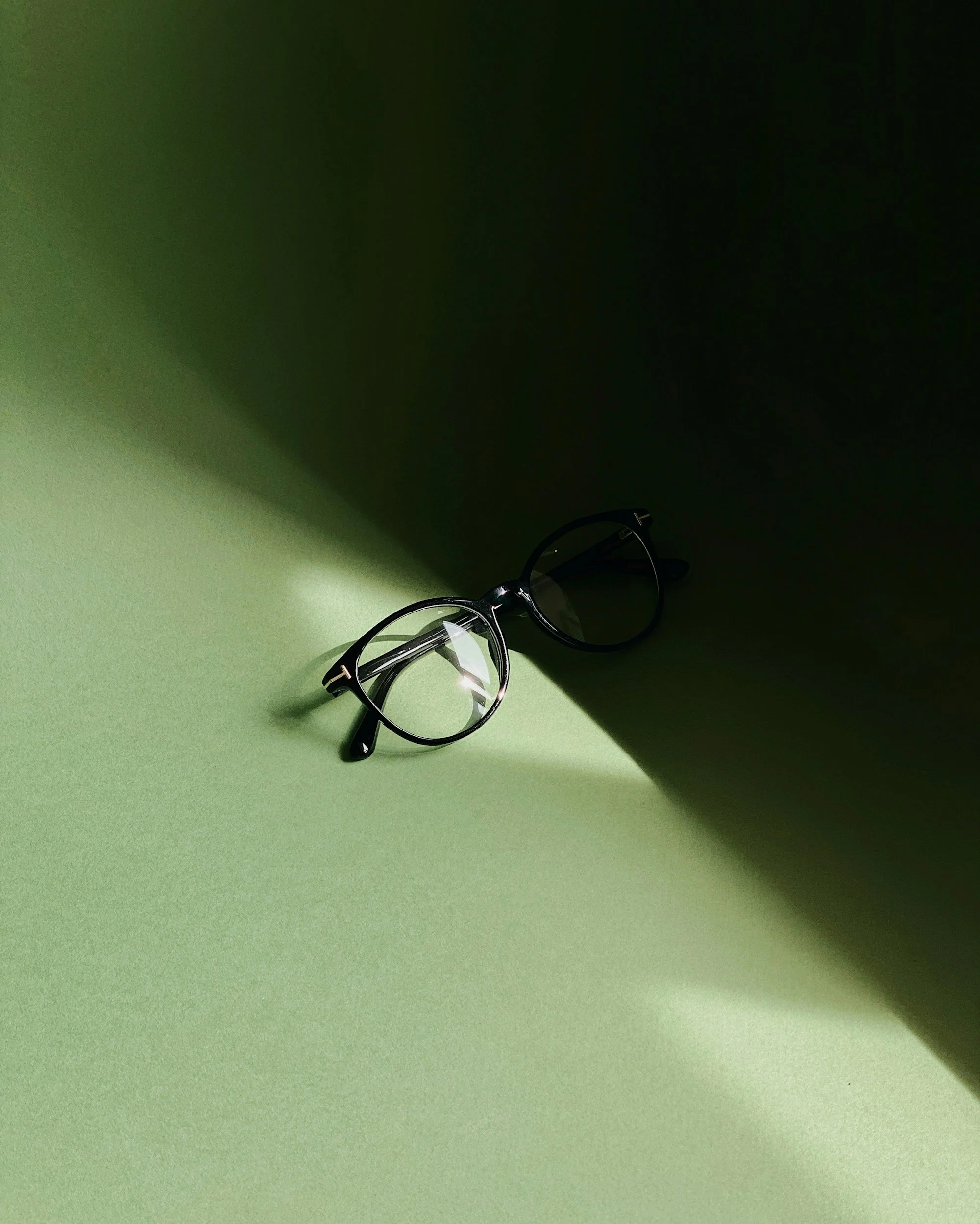 A pair of black eyeglasses with a plastic frame resting on a light green surface, with a shadow cast on the right side of the surface.