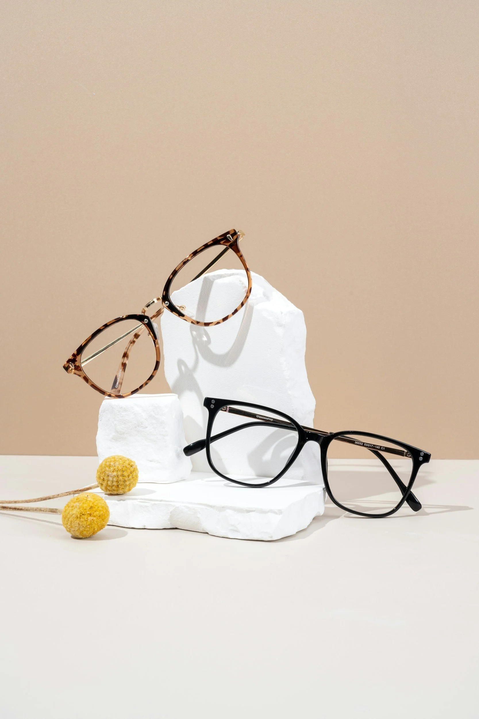 Two pairs of eyeglasses, one with tortoiseshell frames and the other with black frames, displayed on a white textured sculpture against a beige background, with small yellow pom-poms and a brown stick in the foreground.