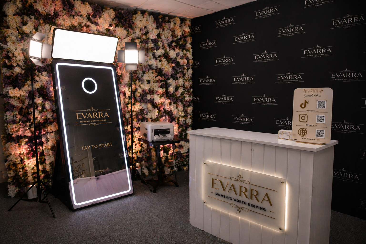 Event setup with a floral backdrop, large digital photo booth screen reading 'EVARRA' and 'TAP TO START', followed by a white counter with 'EVARRA' logo and tagline 'Moments Worth Keeping', a sign with social media icons and QR codes, and professional lighting.