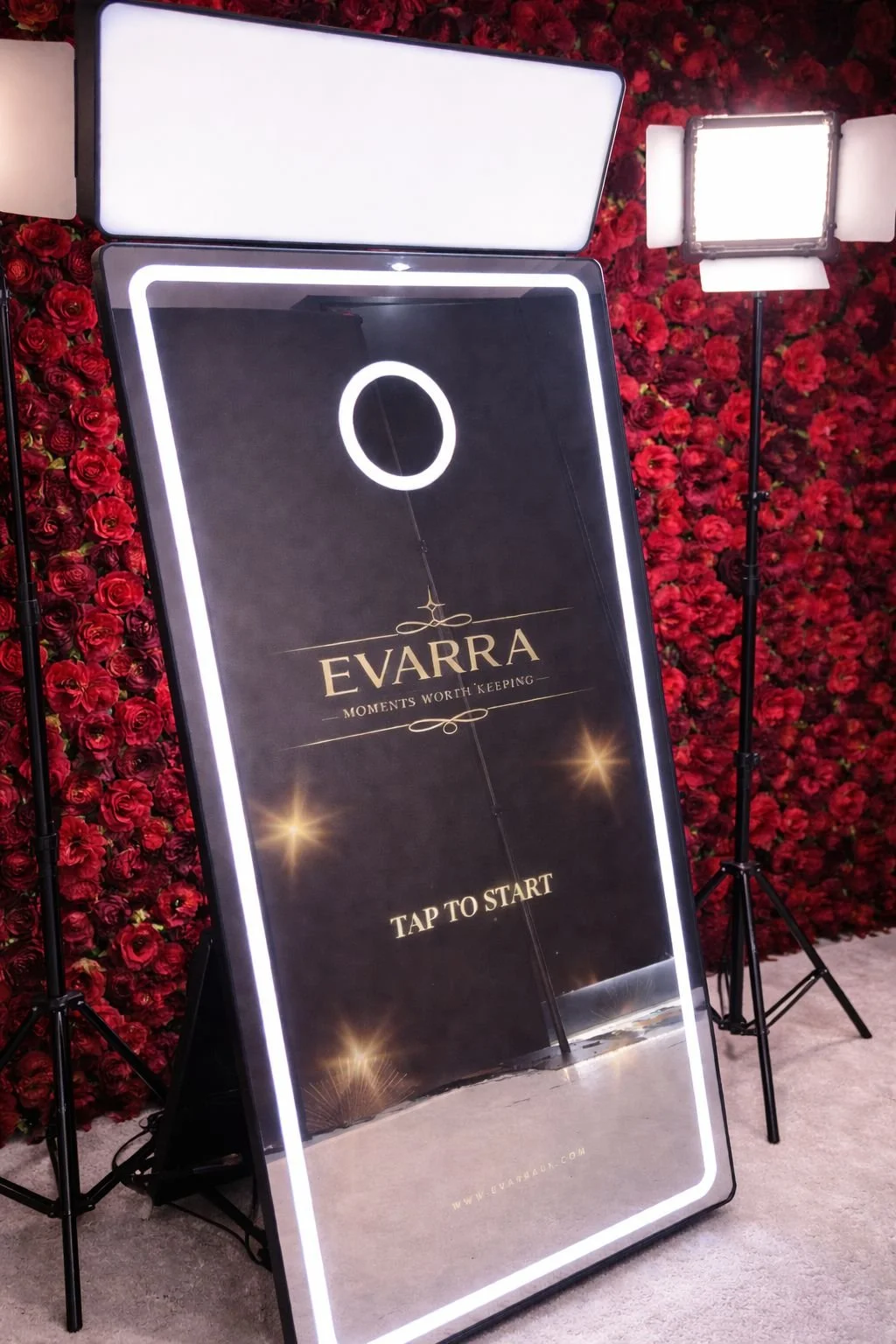 A digital signboard displaying the logo for Evarra with the slogan "Moments Worth Keeping," and a prompt to tap to start, set against a background wall made of red roses with professional lighting equipment around.