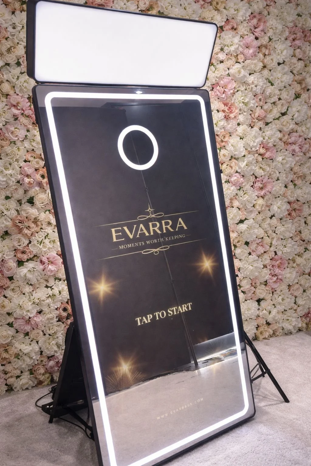 A vertical digital sign with a black background and white border, featuring the brand name EVARRA in gold and the slogan 'Moments Worth Keeping,' with a flower-like design at the top and the words 'Tap to Start' at the bottom, set against a backdrop of pink and white flowers.
