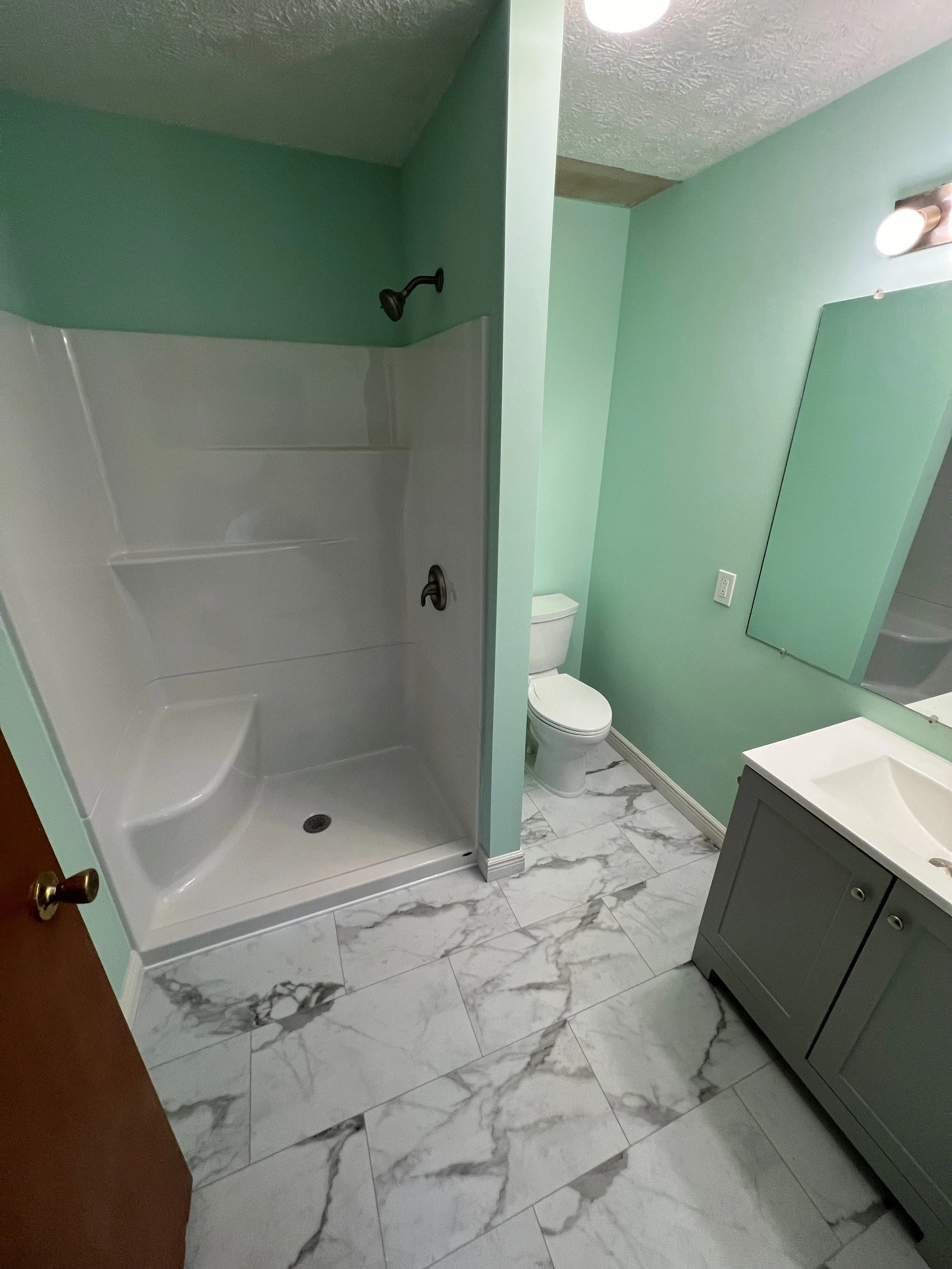 A bathroom with green walls, a shower with black fixtures, a toilet, a mirror, a sink with a gray vanity, and marble-patterned floor tiles.