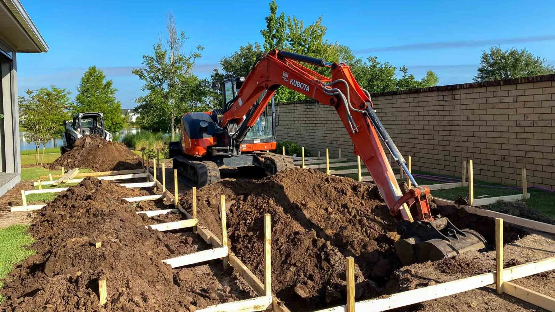 Construction site with excavators digging trenches, wooden frames outline the areas for a building foundation, trees and a brick wall are in the background.