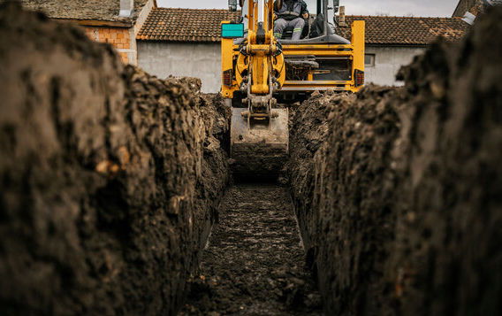 A yellow excavator digging a trench in the ground with a setting of residential buildings in the background.