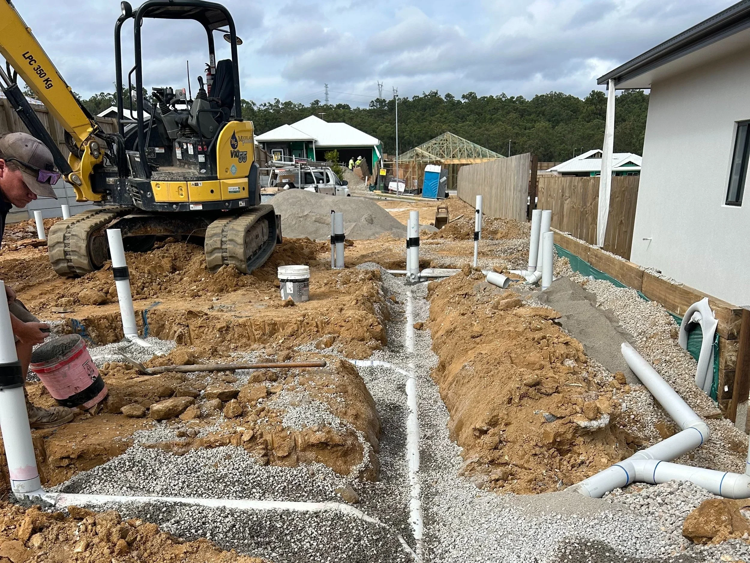 Construction site with a small yellow excavator and visible underground piping being installed in a residential area.