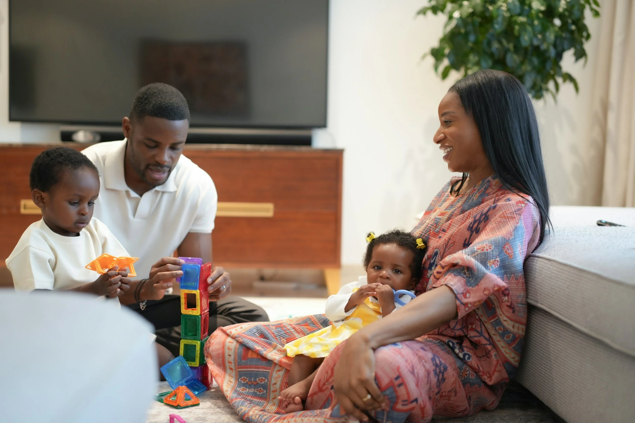 Family sitting on the floor playing with colorful magnetic building blocks, with a mother smiling and a young girl sitting on her lap.