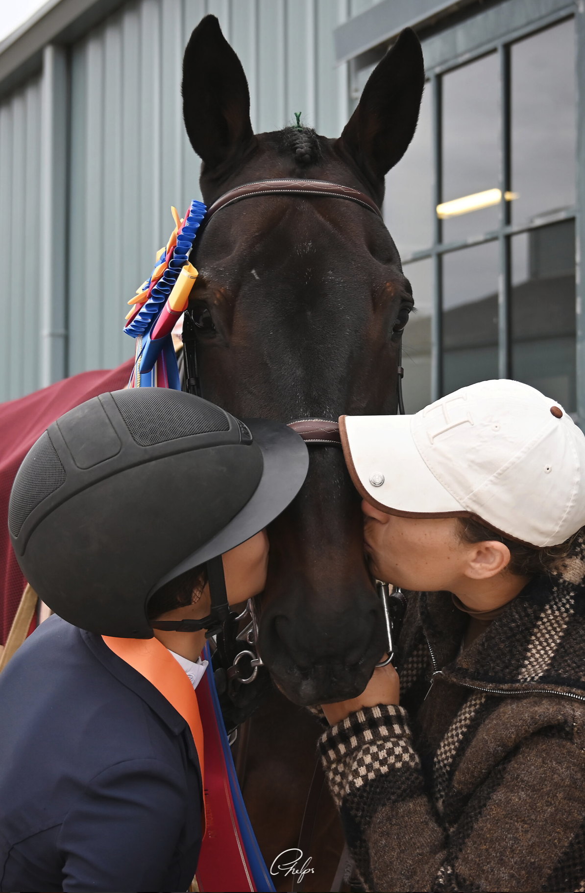 Two women in riding helmets kiss a horse on its nose.