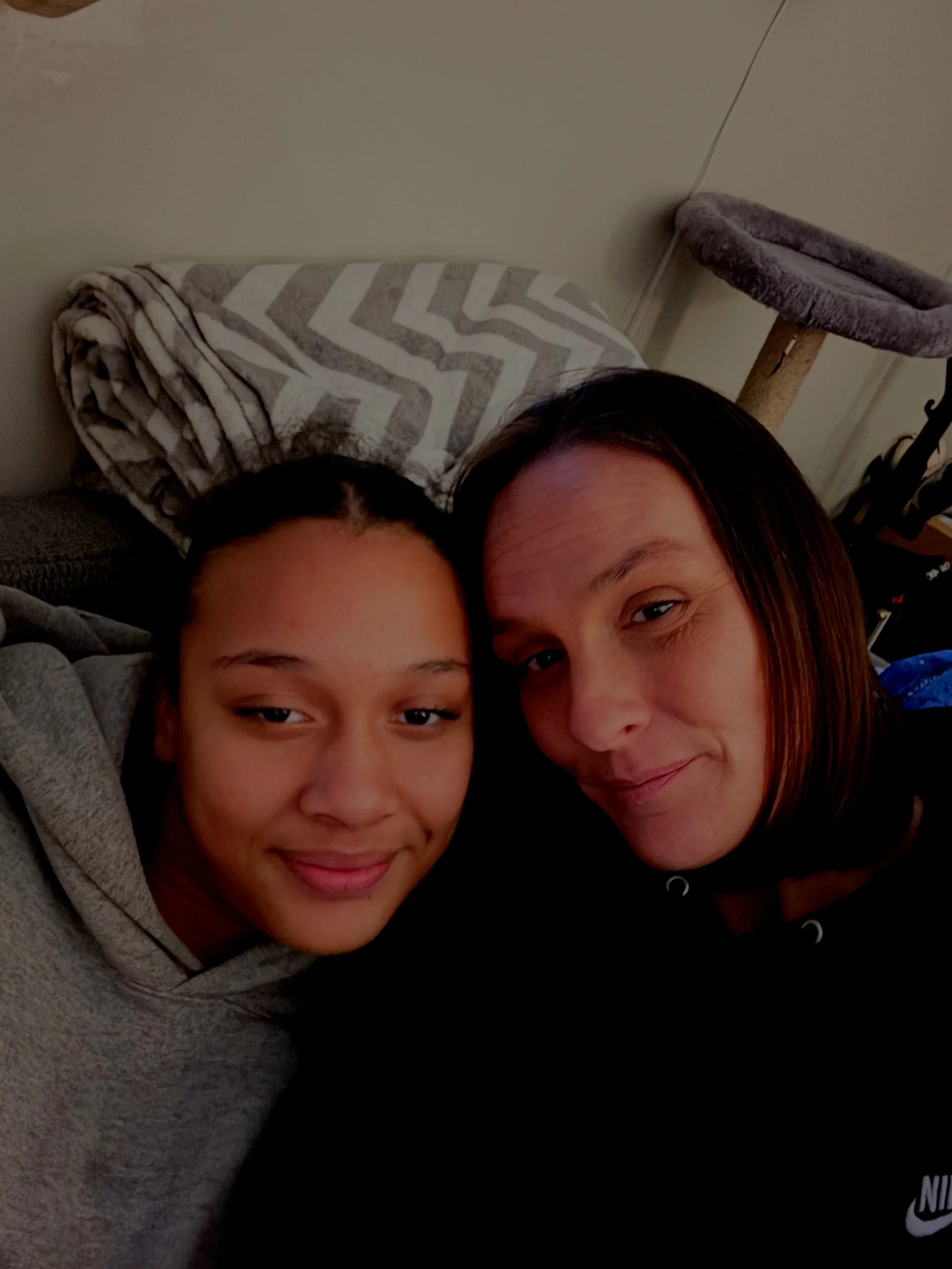 Two women taking a selfie together, smiling, in an indoor setting with a blanket and a cat tree in the background.