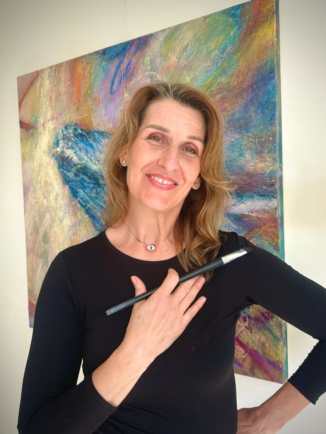 Woman with wavy, shoulder-length hair wearing a black shirt and jewelry, holding a paintbrush against her chest, standing in front of a colorful abstract painting.