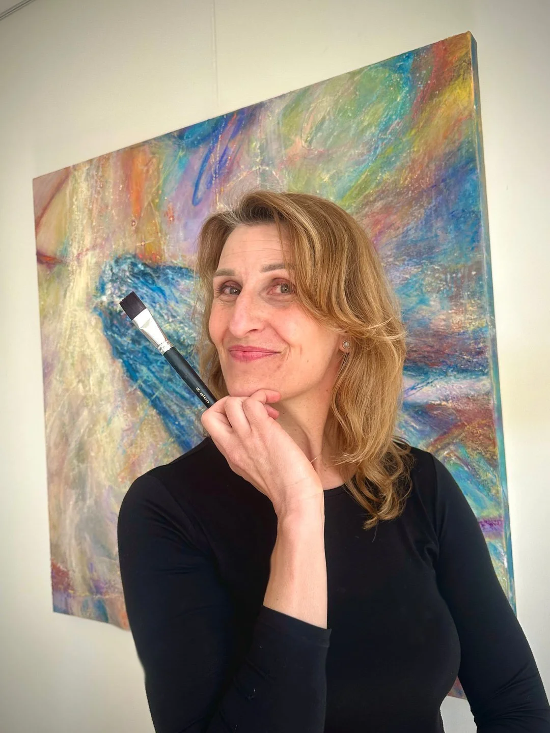 A woman with shoulder-length light brown hair, wearing a black top, is posing in front of an abstract colorful painting. She is holding a paintbrush under her chin, smiling slightly.