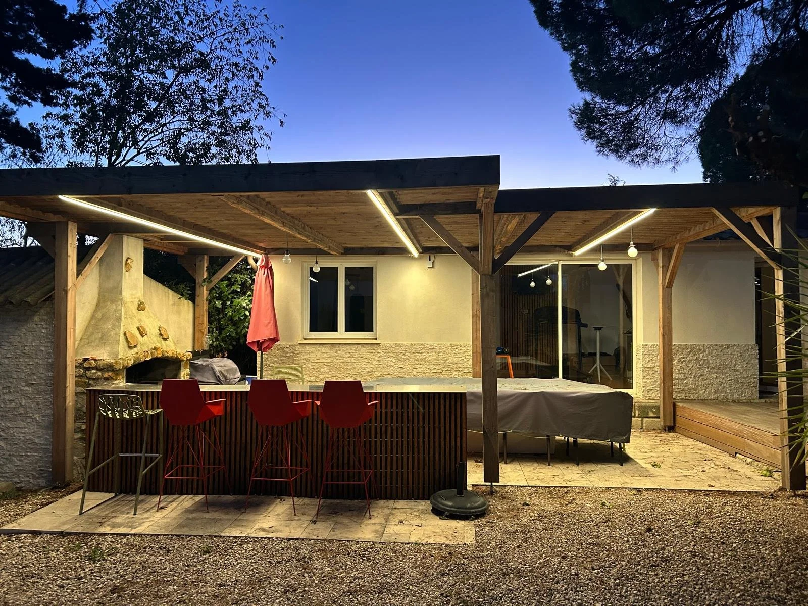 Une terrasse extérieure couverte avec un comptoir de bar rouge, quatre chaises rouges, une table recouverte d'une nappe, une cheminée en pierre, un parasol rose fermé, et un intérieur visible à travers une grande vitre.