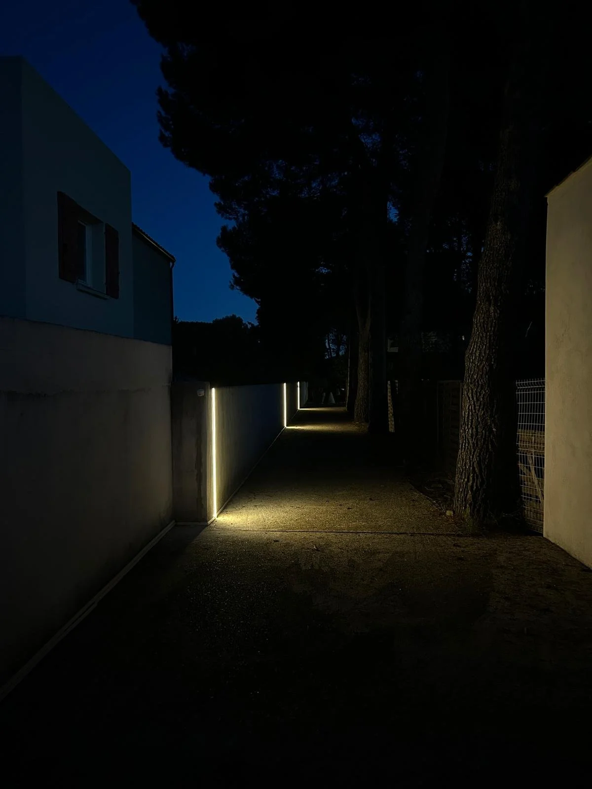 Chemin éclairé par des lumières LED le long d'un mur dans une rue nocturne, avec des arbres en arrière-plan.