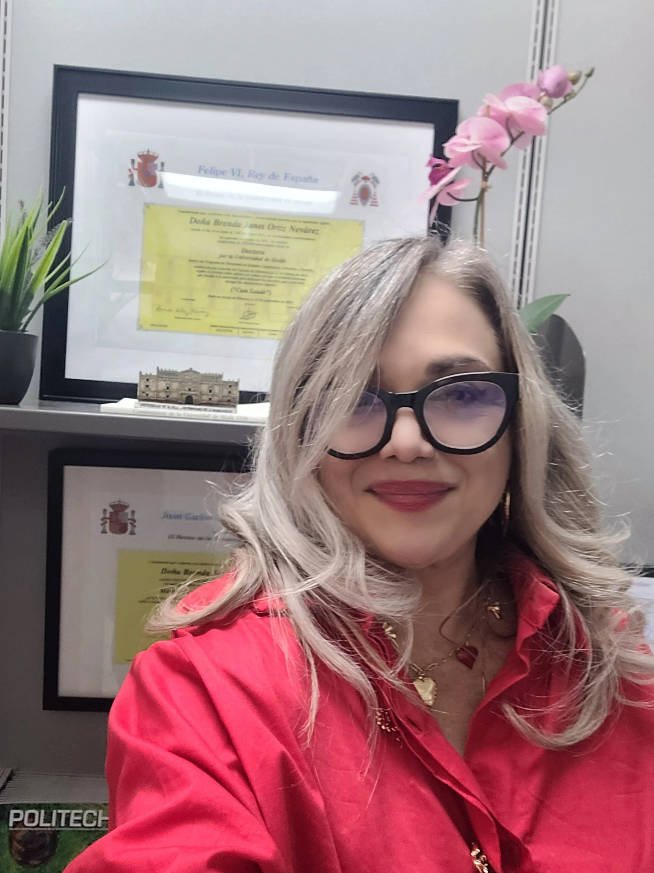 A woman with blonde hair, wearing large glasses and a red shirt, taking a selfie in front of framed certificates and decorations on a wall.