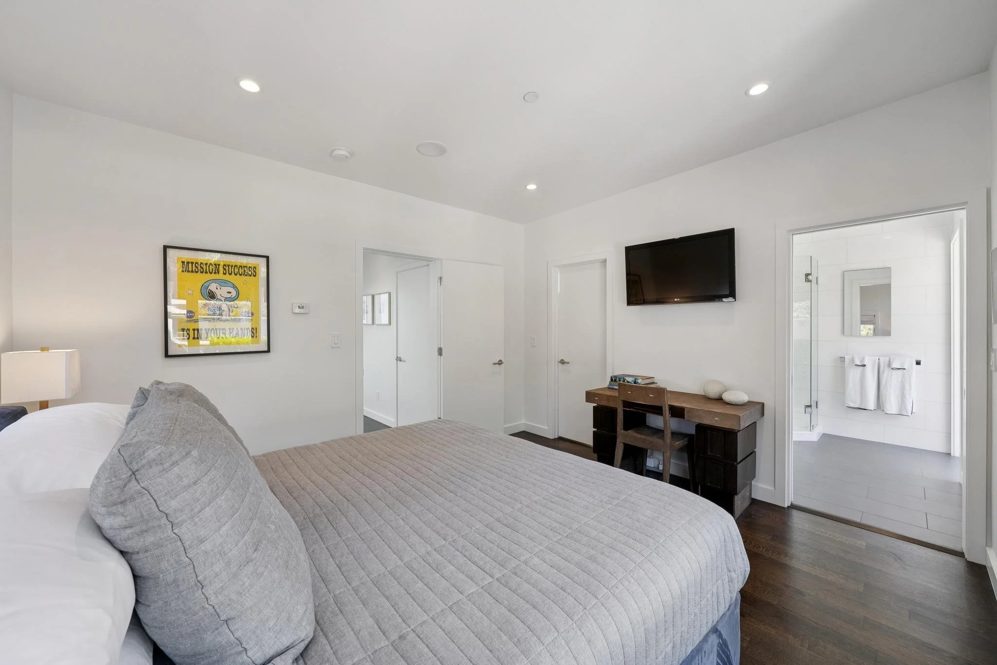 Bedroom with a bed, a framed artwork on the wall, a wall-mounted TV, a small desk with decorative objects, and a door opening to a bathroom.