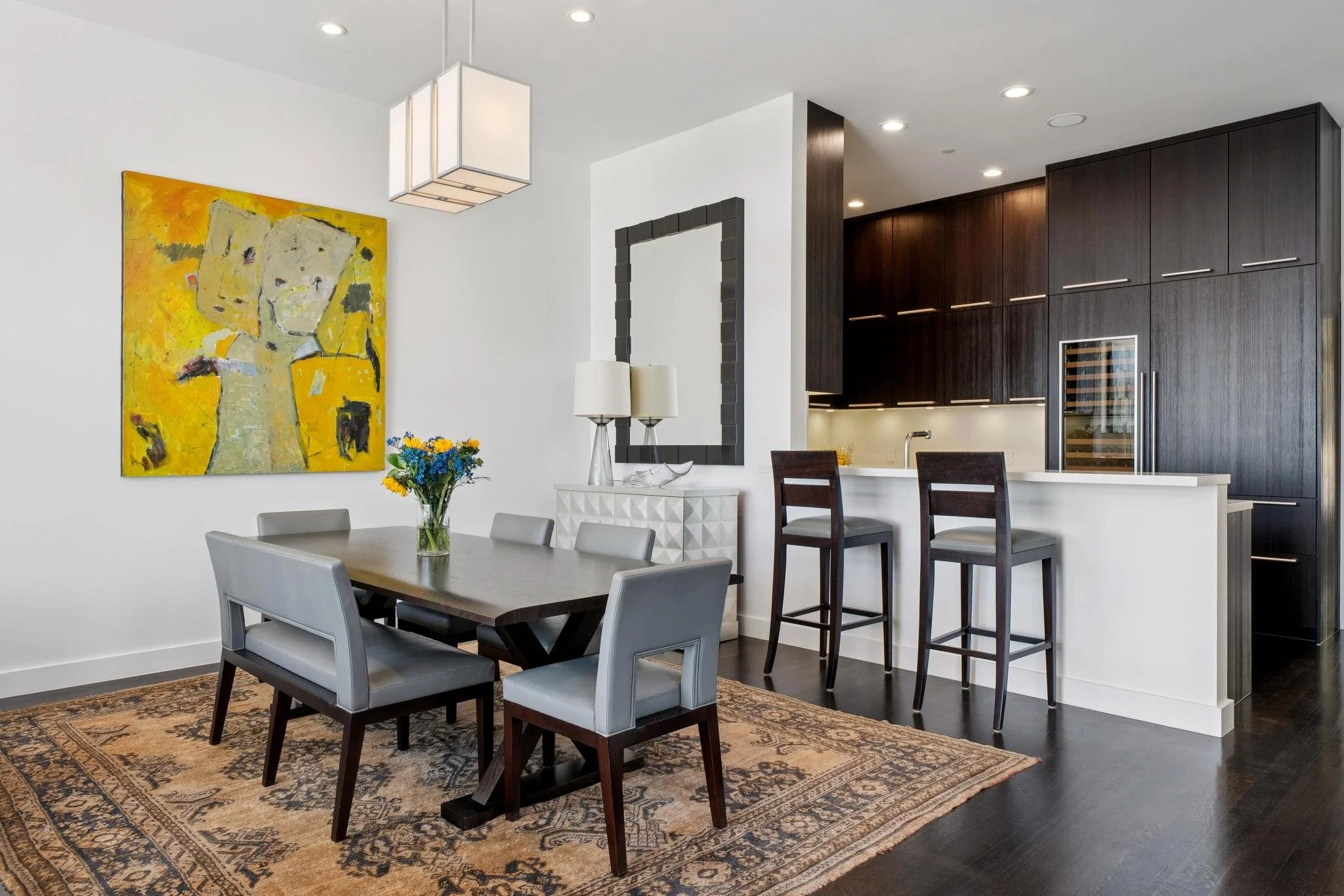 Modern dining area with six light gray upholstered chairs around a dark wood table, a vase with yellow and blue flowers, a large colorful abstract painting on the white wall, a decorative mirror, and a white sideboard with lamps, next to a kitchen wi