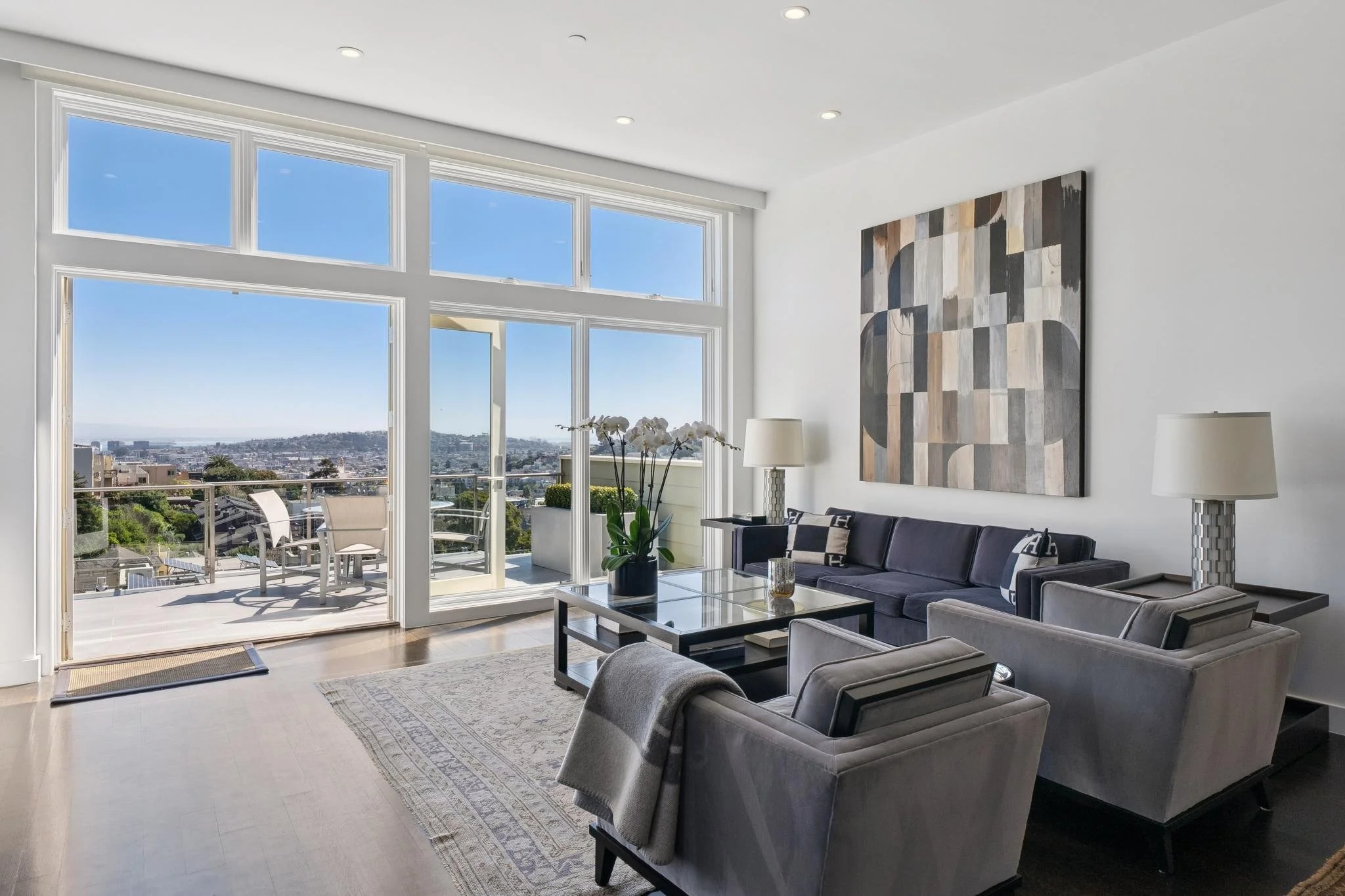 Bright living room with large windows and a view of an outdoor balcony with patio furniture. Contains gray sofas, armchairs, a glass coffee table with a potted orchid, and wall art. Two table lamps on side tables.