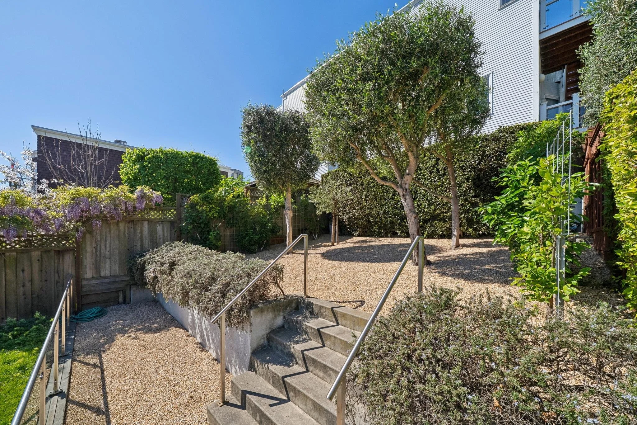 A backyard garden with a concrete staircase, metal handrails, trees, shrubs, a wooden fence, and a house with white siding under a clear blue sky.