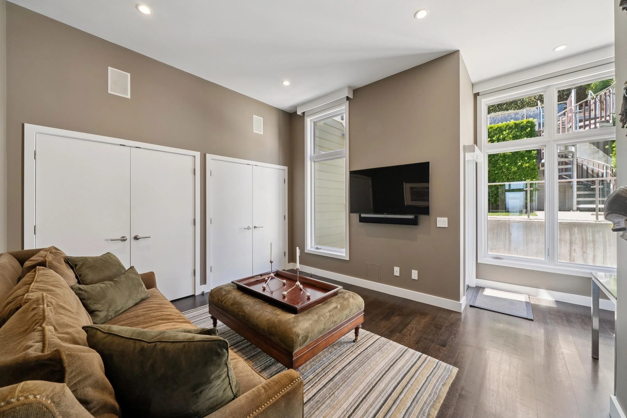 Living room with a brown couch, a patterned area rug, a wall-mounted TV, large windows showing greenery outside, and white double closet doors.