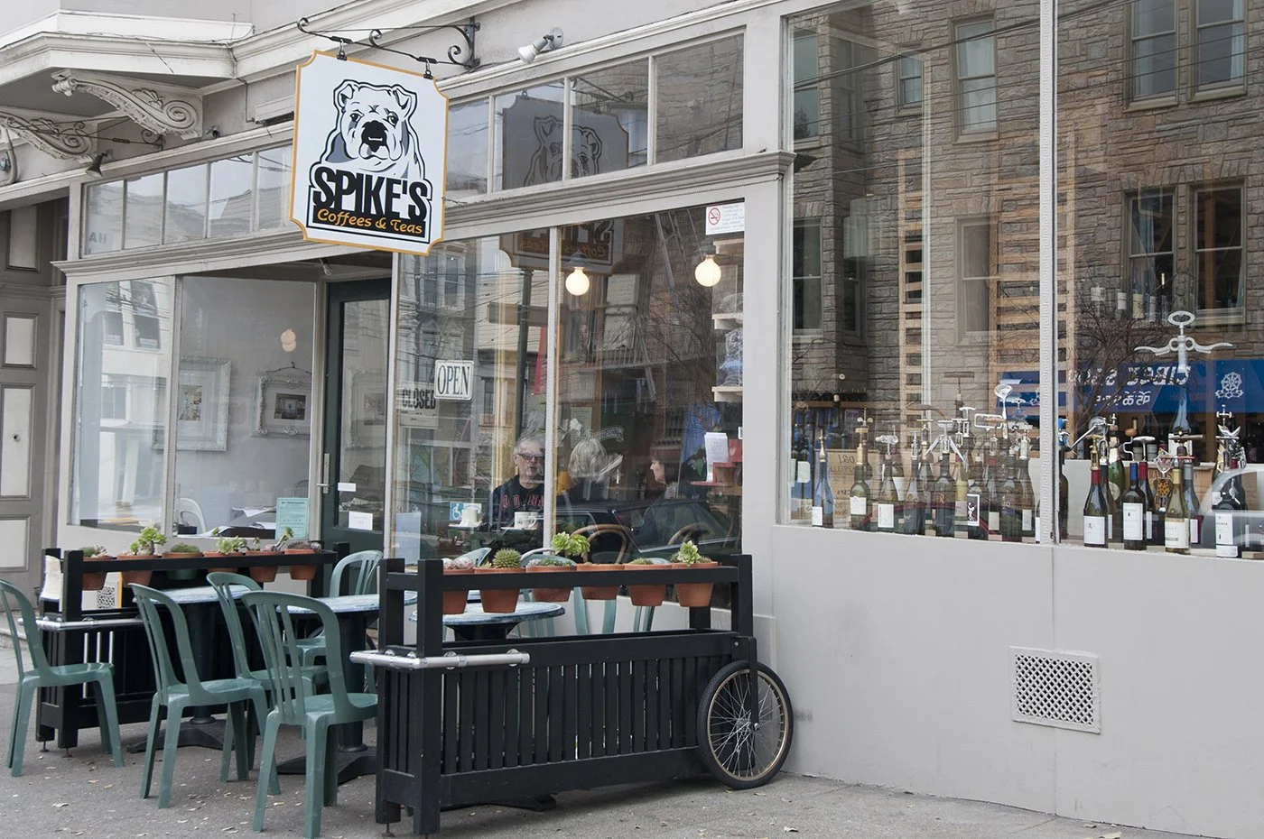 Exterior view of Spike's Coffees & Teas shop with outdoor seating, potted plants, and a bicycle leaning against a black fence. The shop has large glass windows, and insideoppers are visible. A sign above the shop shows a dog logo.