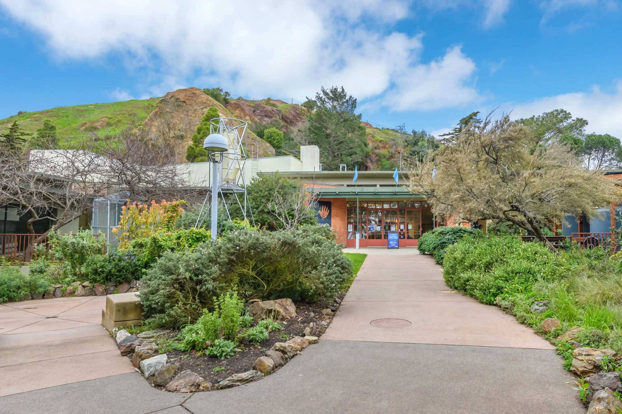 Entrance to a public building, possibly a museum or visitor center, with a pathway, landscaped garden, and a hill with trees and rocks in the background.