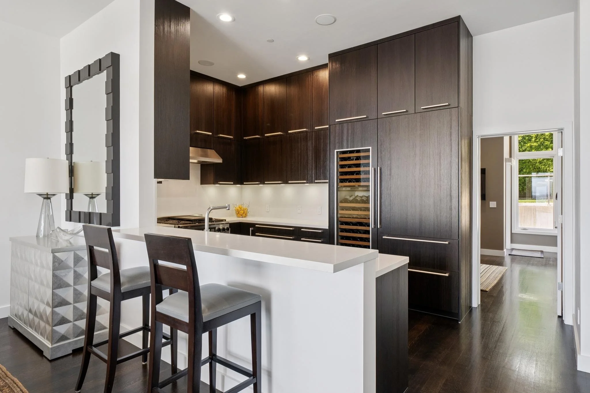 Modern kitchen with dark wood cabinets, white countertops, and a built-in wine cooler, adjacent to a white dining area with black chairs and a decorative mirror.