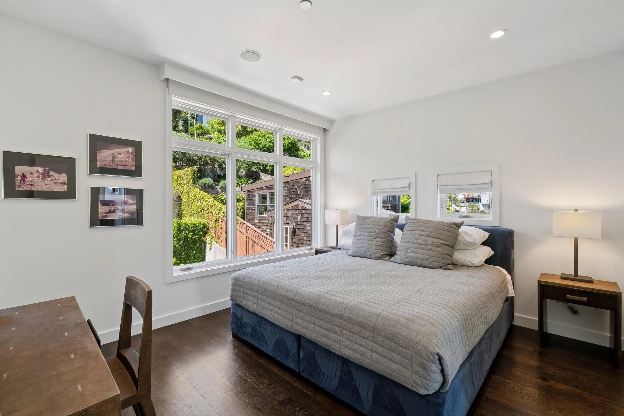 Bright bedroom with large window view of greenery, white walls, a bed with gray pillows, and wooden furniture.