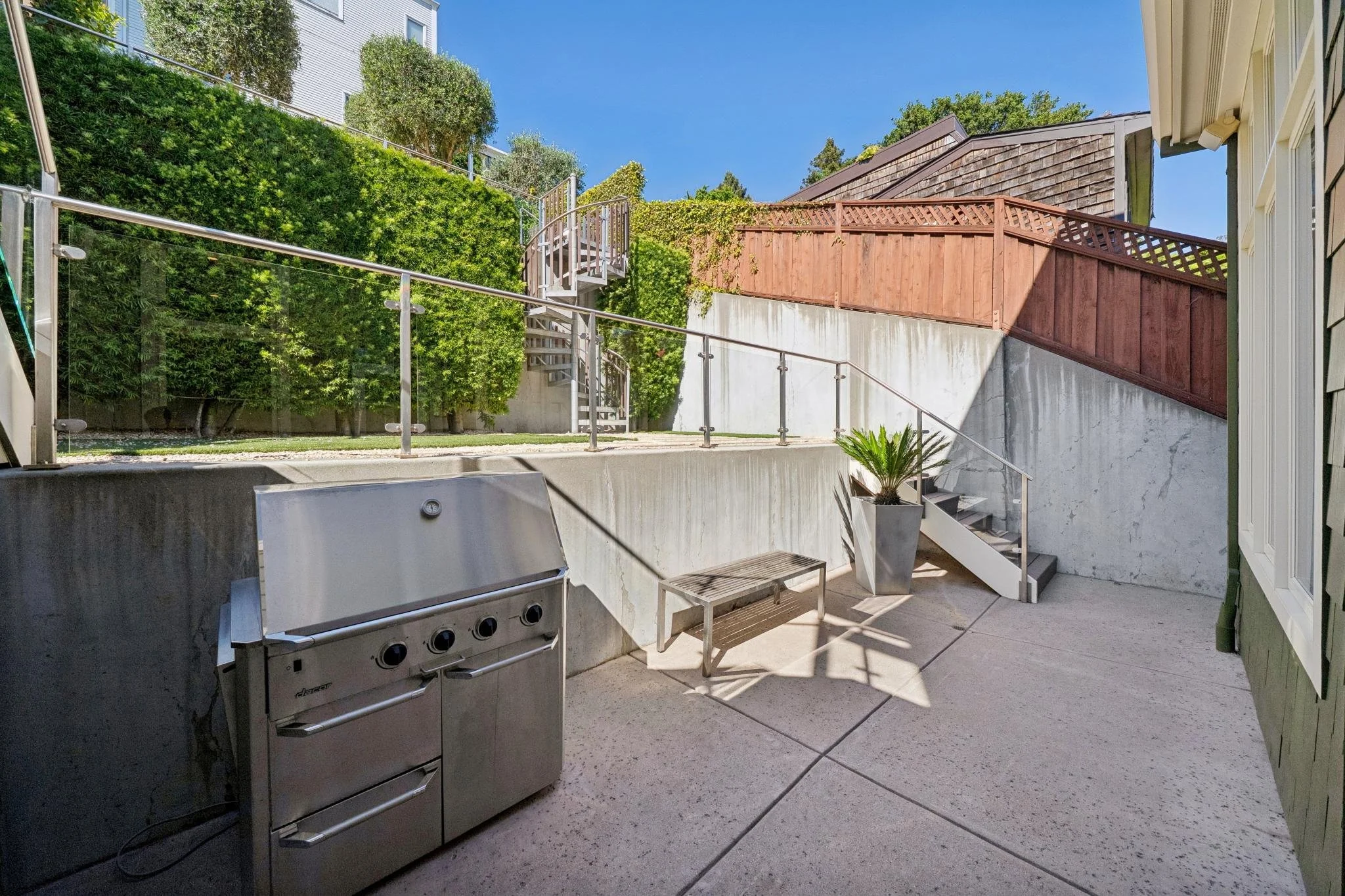 Outdoor patio with a stainless steel grill, a bench, a large potted plant, concrete stairs with metal railing, green bushes, and a wooden fence under a clear blue sky.