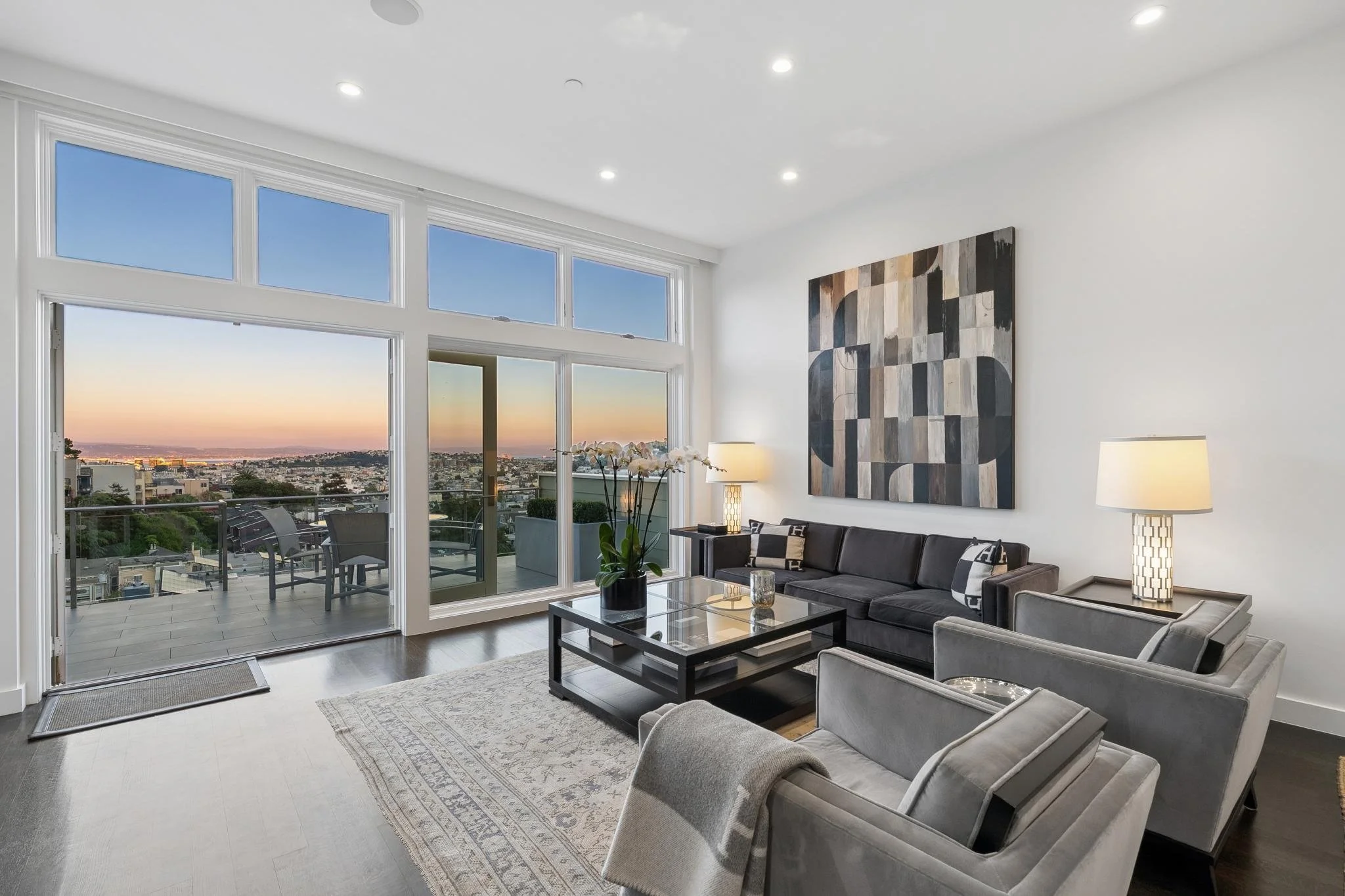 Modern living room with large glass doors leading to balcony, city views at sunset, black and gray furniture, abstract wall art, and ambient lighting.
